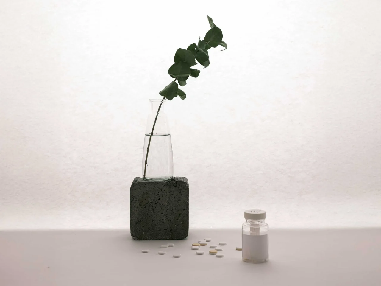 Assorted white and beige tablets scattered on a white surface next to a small glass bottle and a dark brick-shaped block on the left.