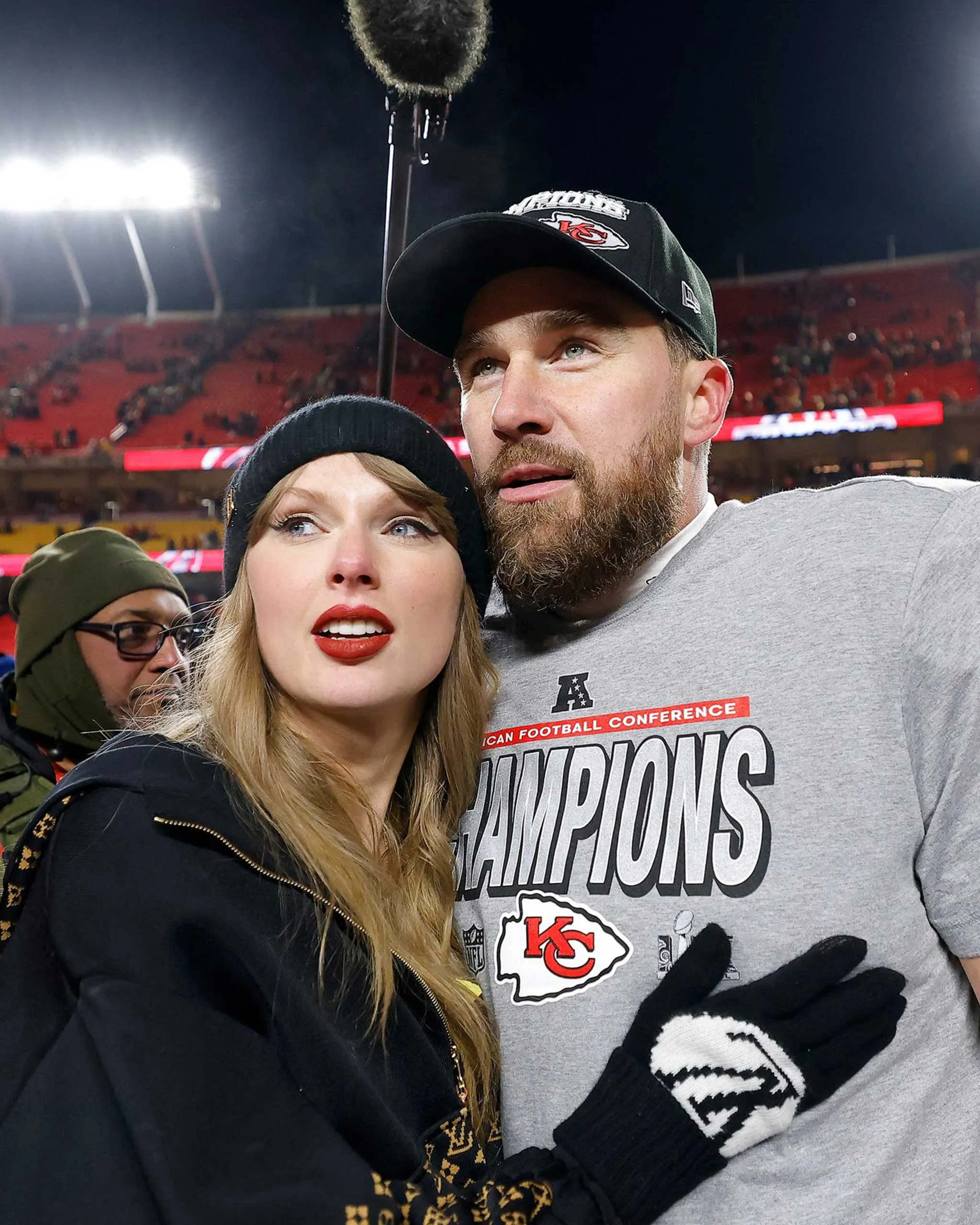 A couple posing for a selfie at a night football stadium, she wears a black beanie and lipstick, he wears a cap and gray champions shirt with a Kansas City logo.