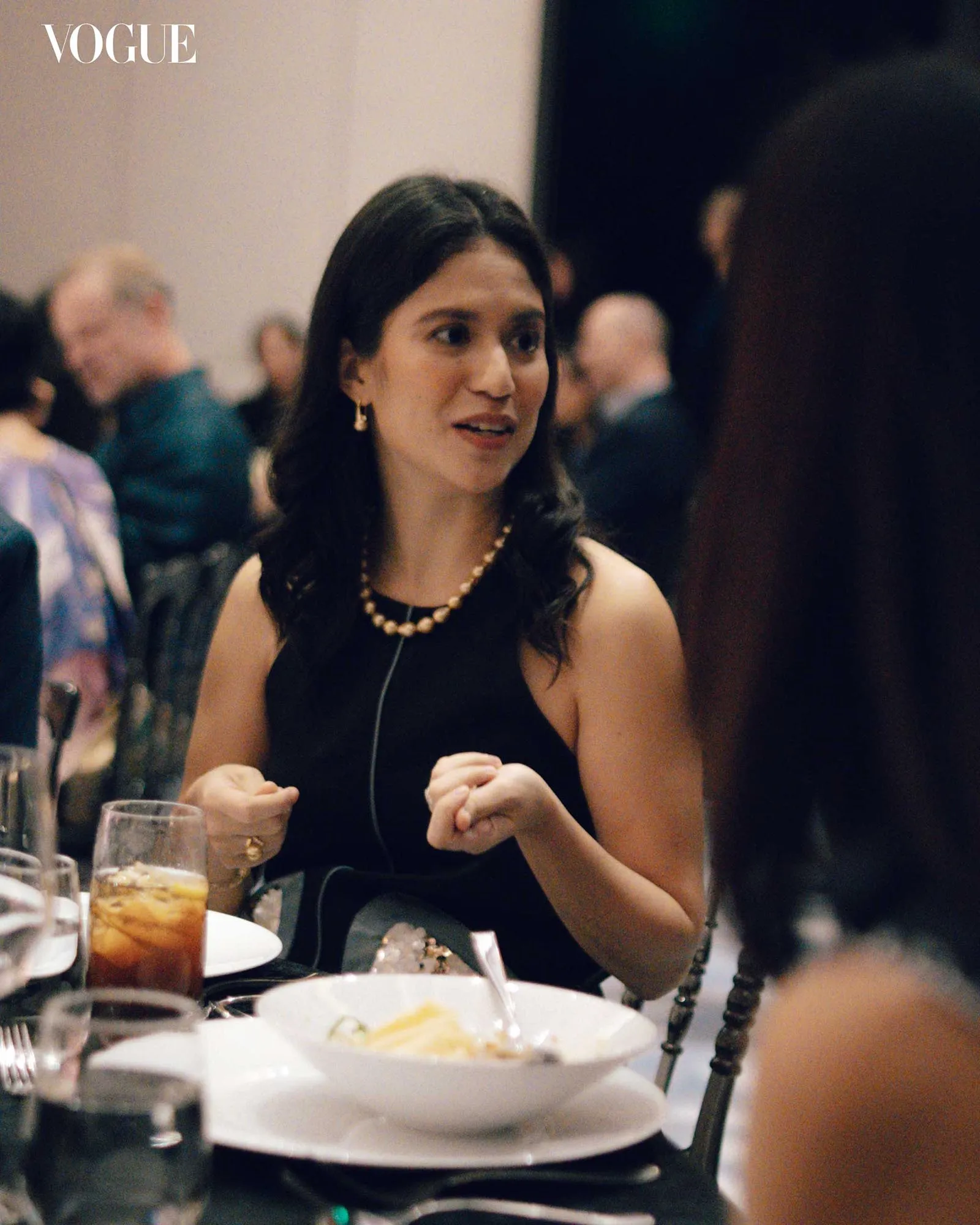 Woman in a black dress with pearl necklace seated at a dining table, speaking in a social event with drinks and plates in view.