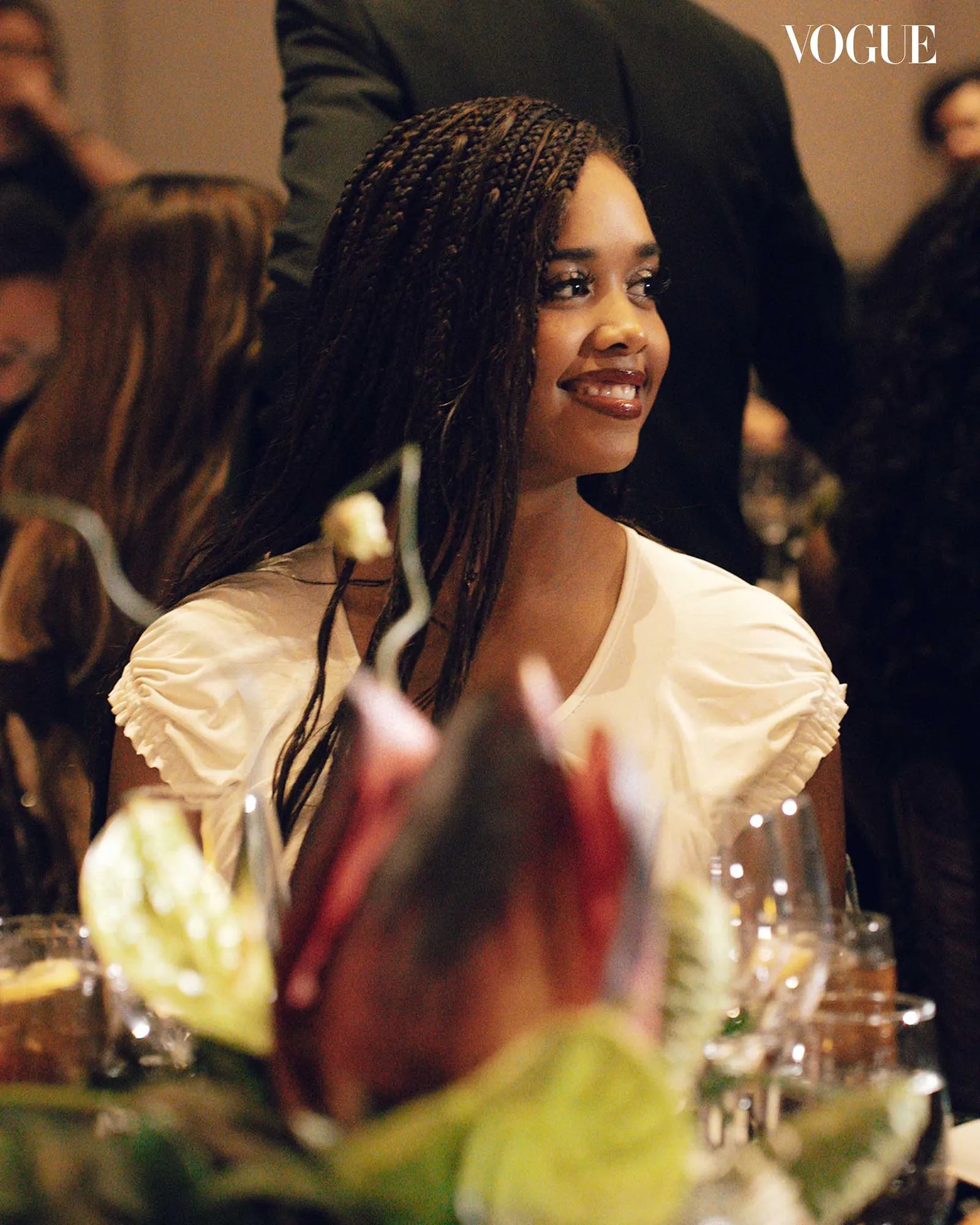 Smiling woman with braided hair seated at a dimly lit table with wine glasses; Vogue logo in the corner.