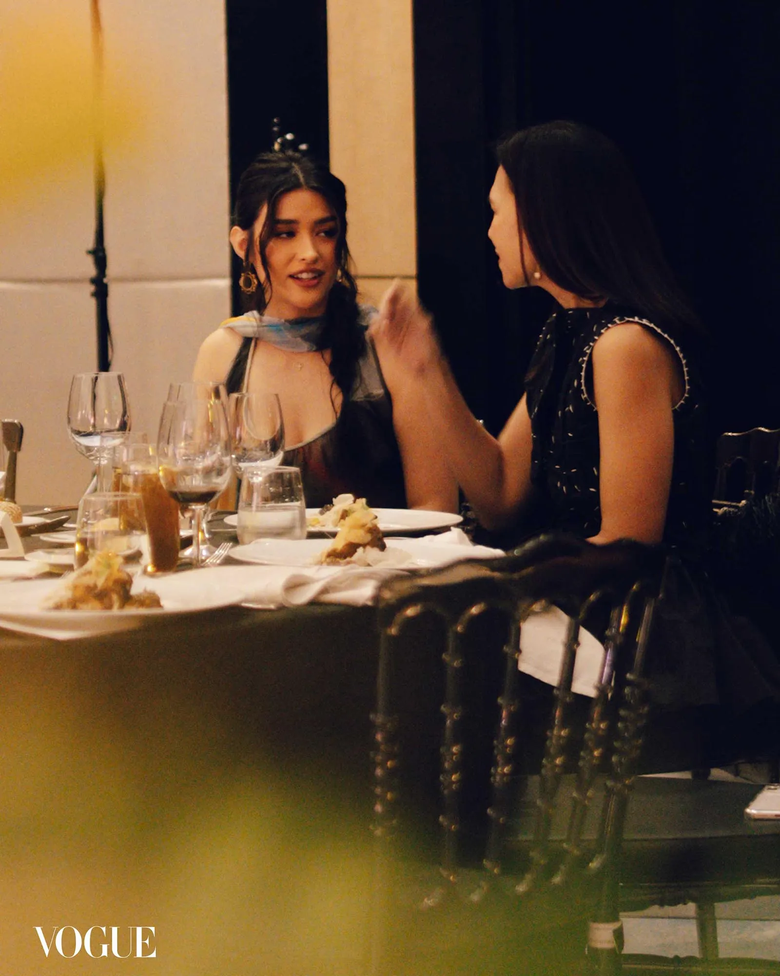 Two women talk across a dining table with wine glasses and plates; Vogue watermark bottom left.