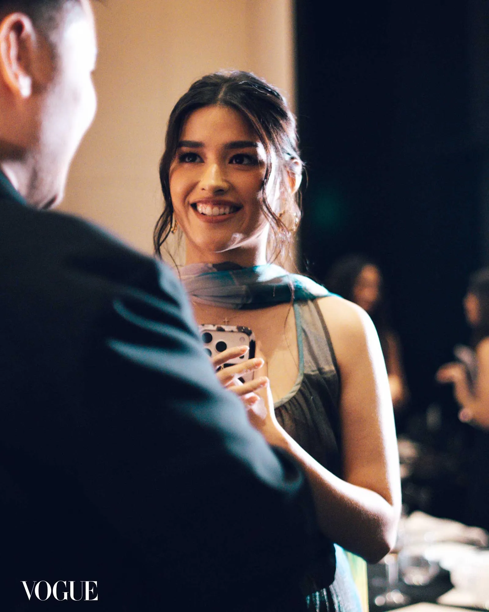 Smiling woman in a sleeveless dress chats with a man at a social event; Vogue logo bottom-left.