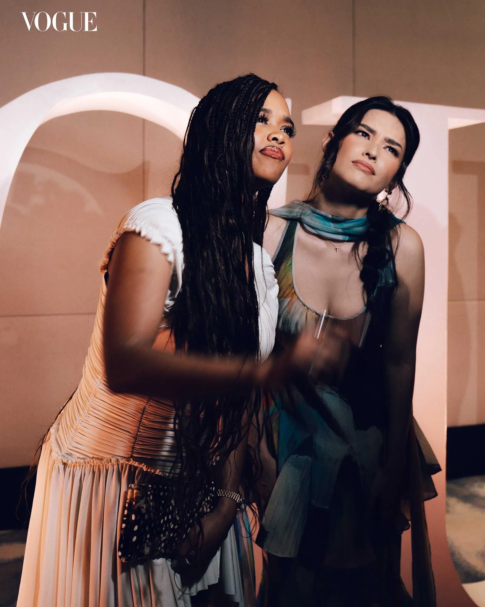 Two women posing together at a Vogue event, wearing colorful evening dresses and looking upward.