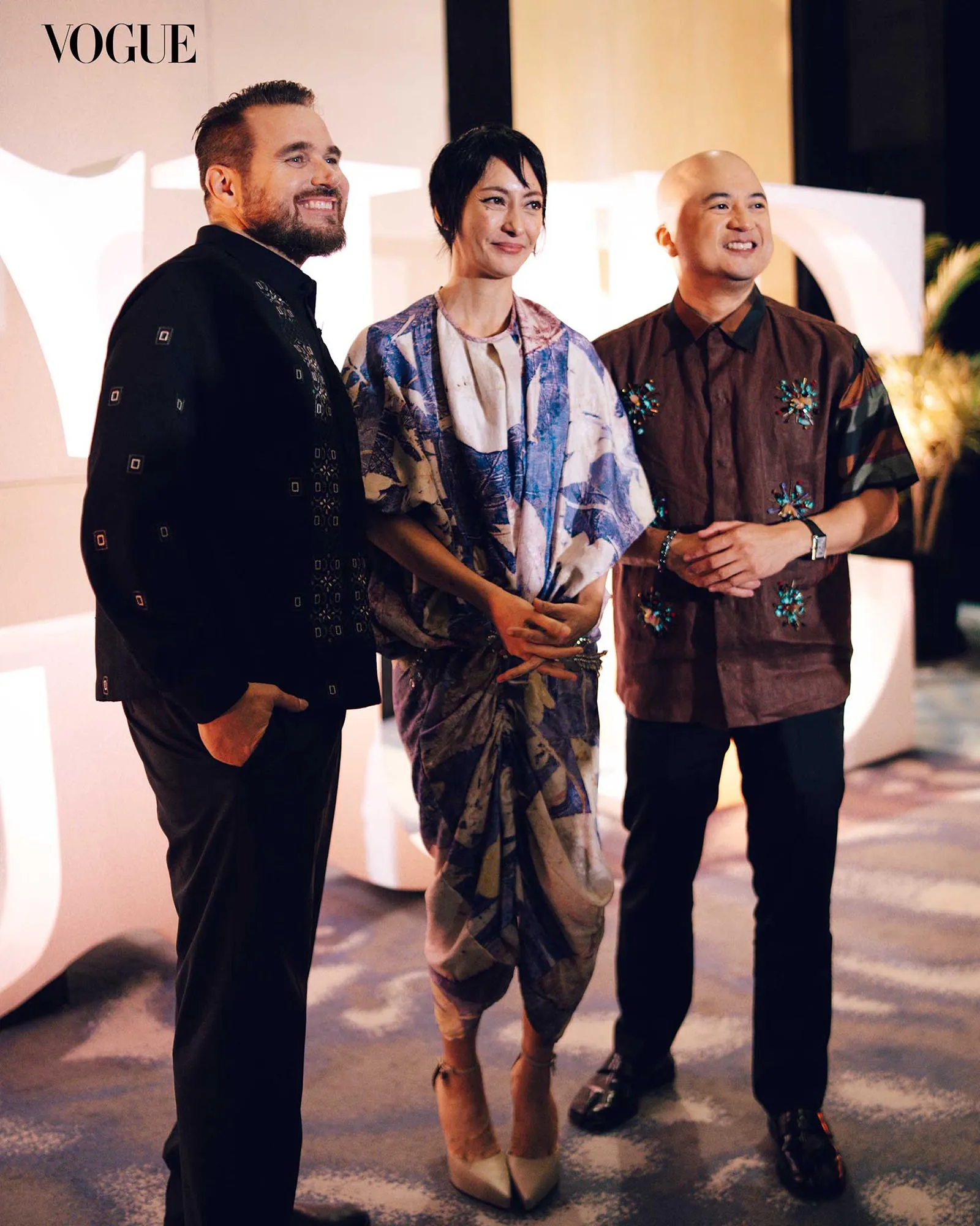 Three people stand together at a Vogue event, smiling for the camera in stylish outfits.