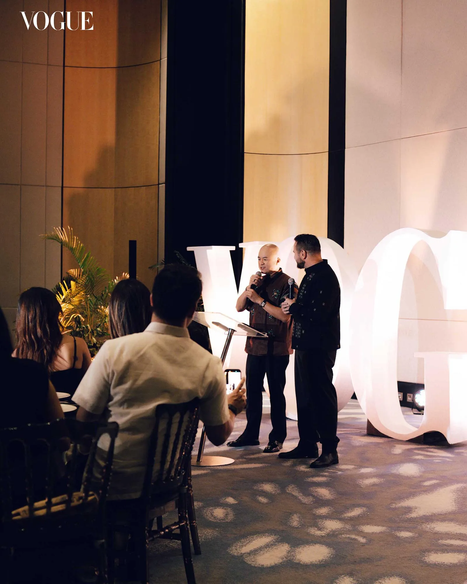 Two men speak into microphones on a stage at a Vogue event, with large white letters behind them and an audience in front.