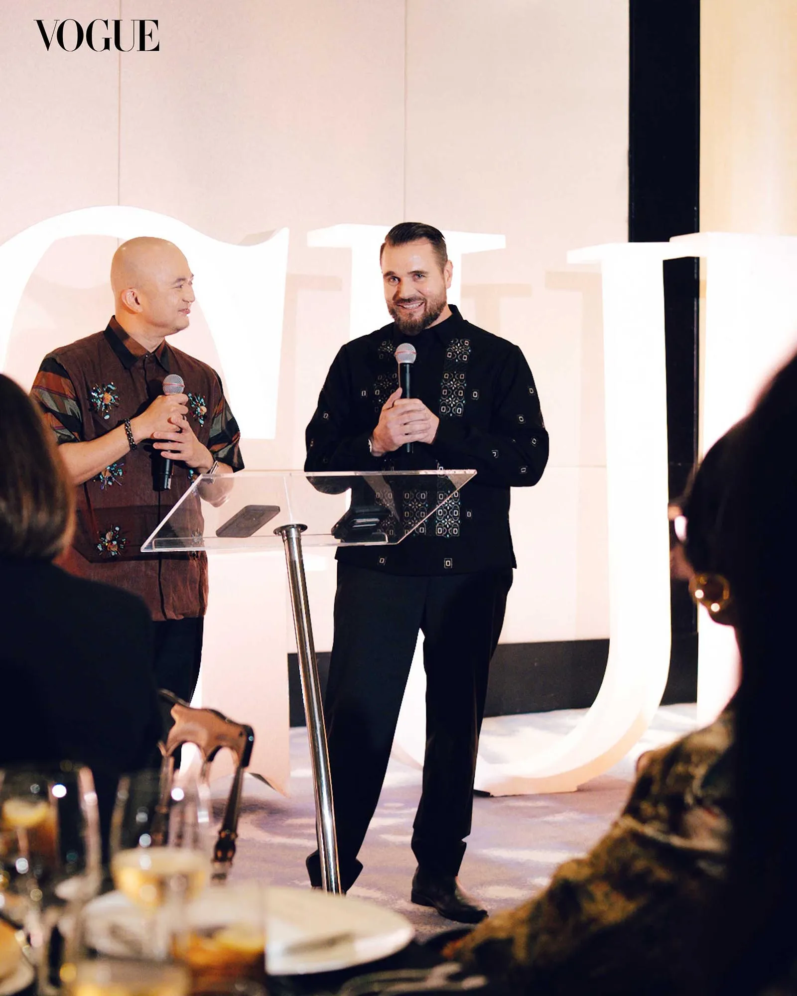 Two men in patterned shirts speak into handheld microphones at a Vogue event, standing behind clear lecterns on a lit stage.