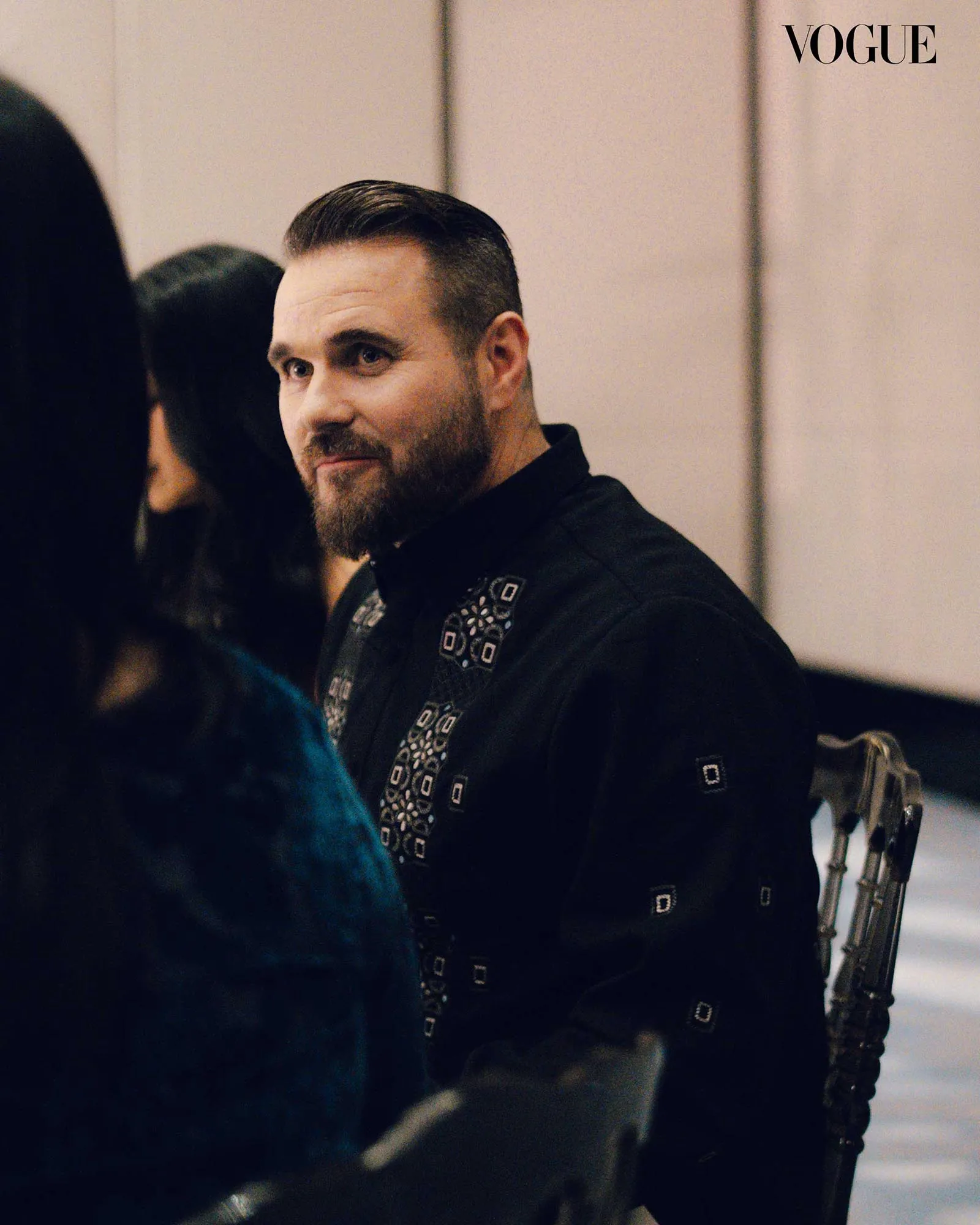 Man with short hair and trimmed beard in a black patterned shirt, gazing to the left in a neutral indoor setting.