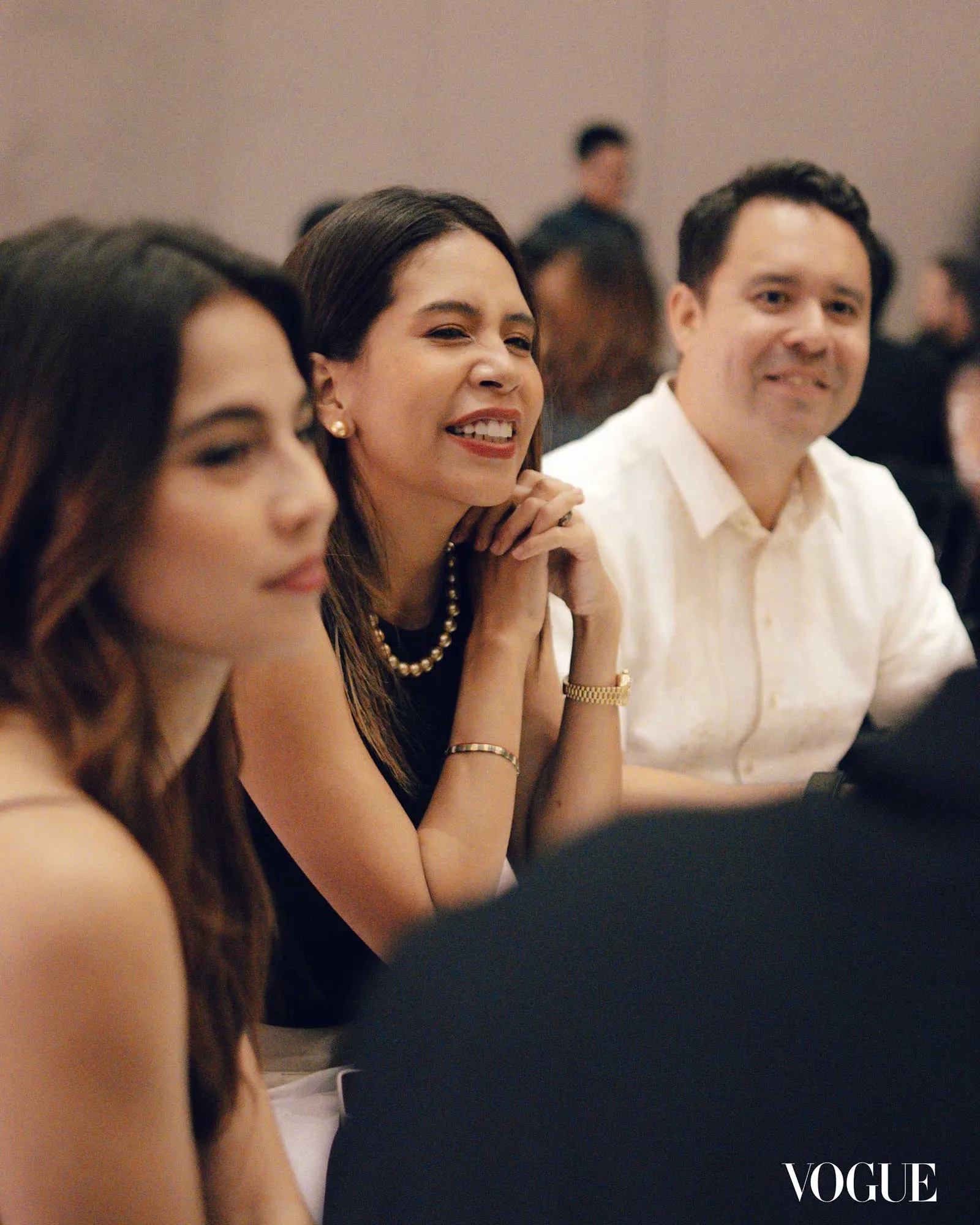 Smiling woman with a pearl necklace at a formal event, others blurred in the background; Vogue logo in bottom-right corner.