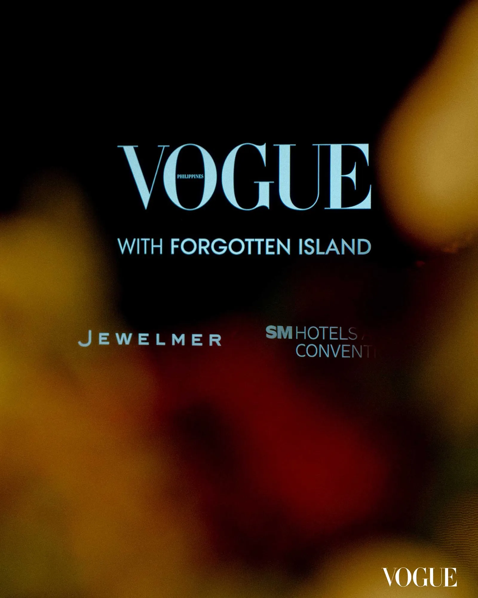 Blurred warm-toned background with a large blue VOGUE logo and the subtitle 'WITH FORGOTTEN ISLAND' on a dark page, resembling a magazine cover.