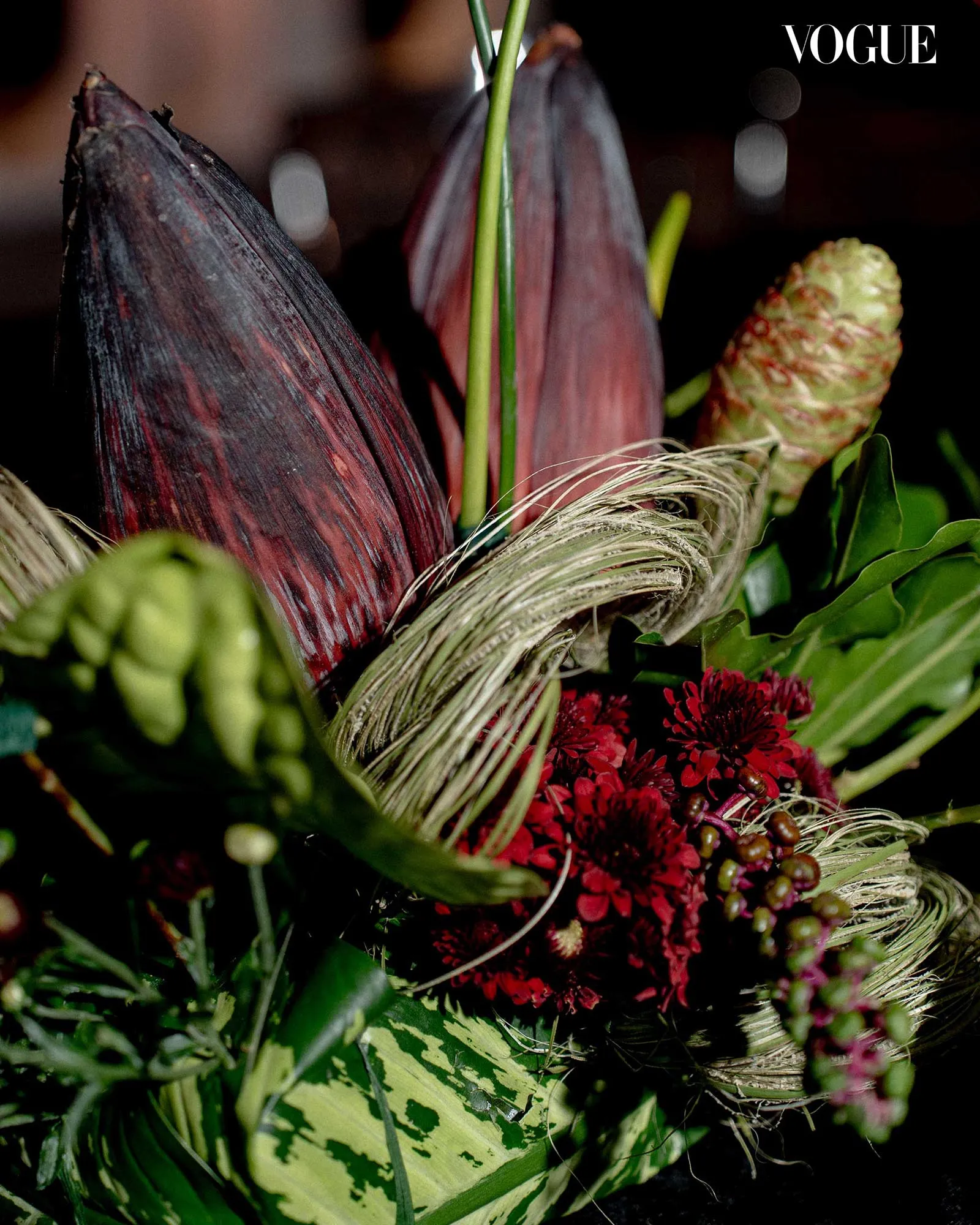Close-up of an exotic floral arrangement with dark purple leaves, green stems, dried grasses, pinecone, and red blossoms; Vogue logo in the corner.