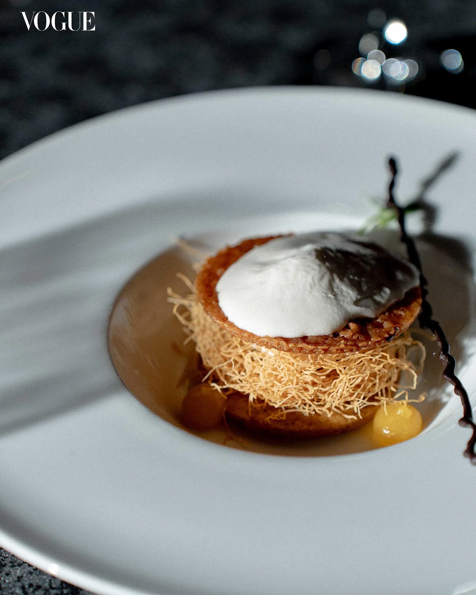 Close-up of a layered dessert with a crisp kataifi nest, white cream dollop, and chocolate drizzle on a white plate; Vogue logo in corner.