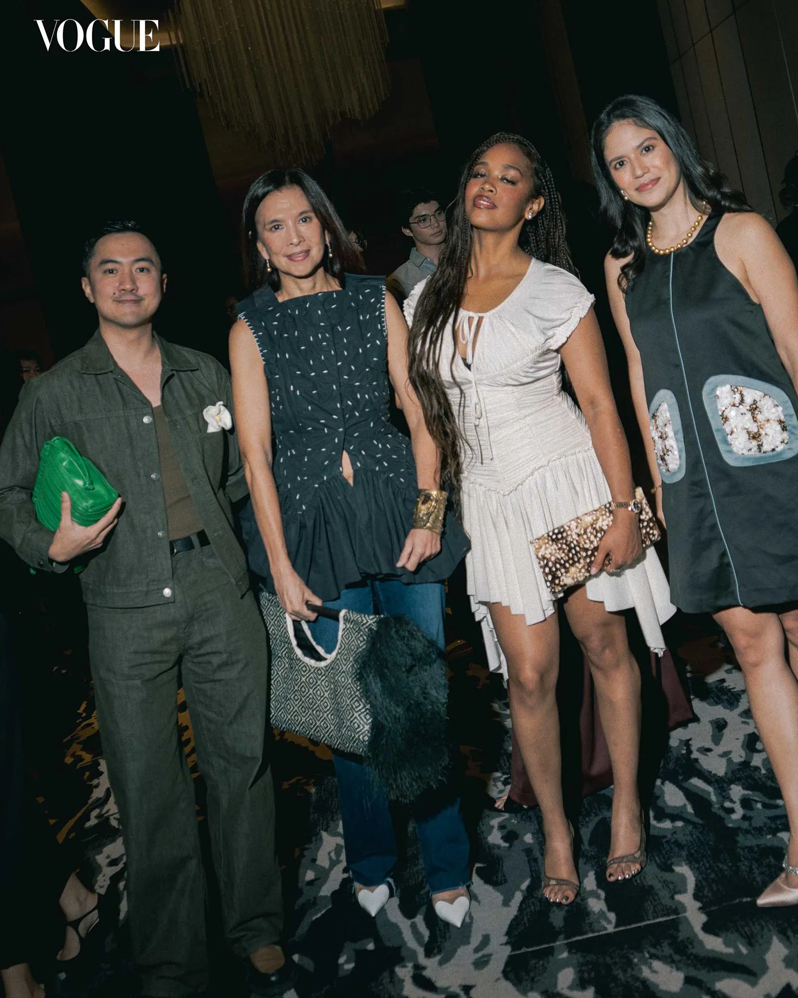 Vogue logo in upper left; four stylish adults pose for a fashion-event photo, each wearing chic outfits and accessories.