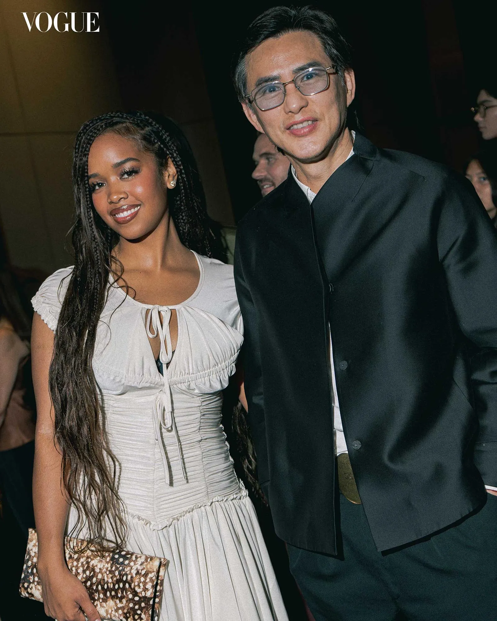 Woman with long braids in a light gray dress posing beside a man in a dark suit at a Vogue event; she holds a patterned clutch.