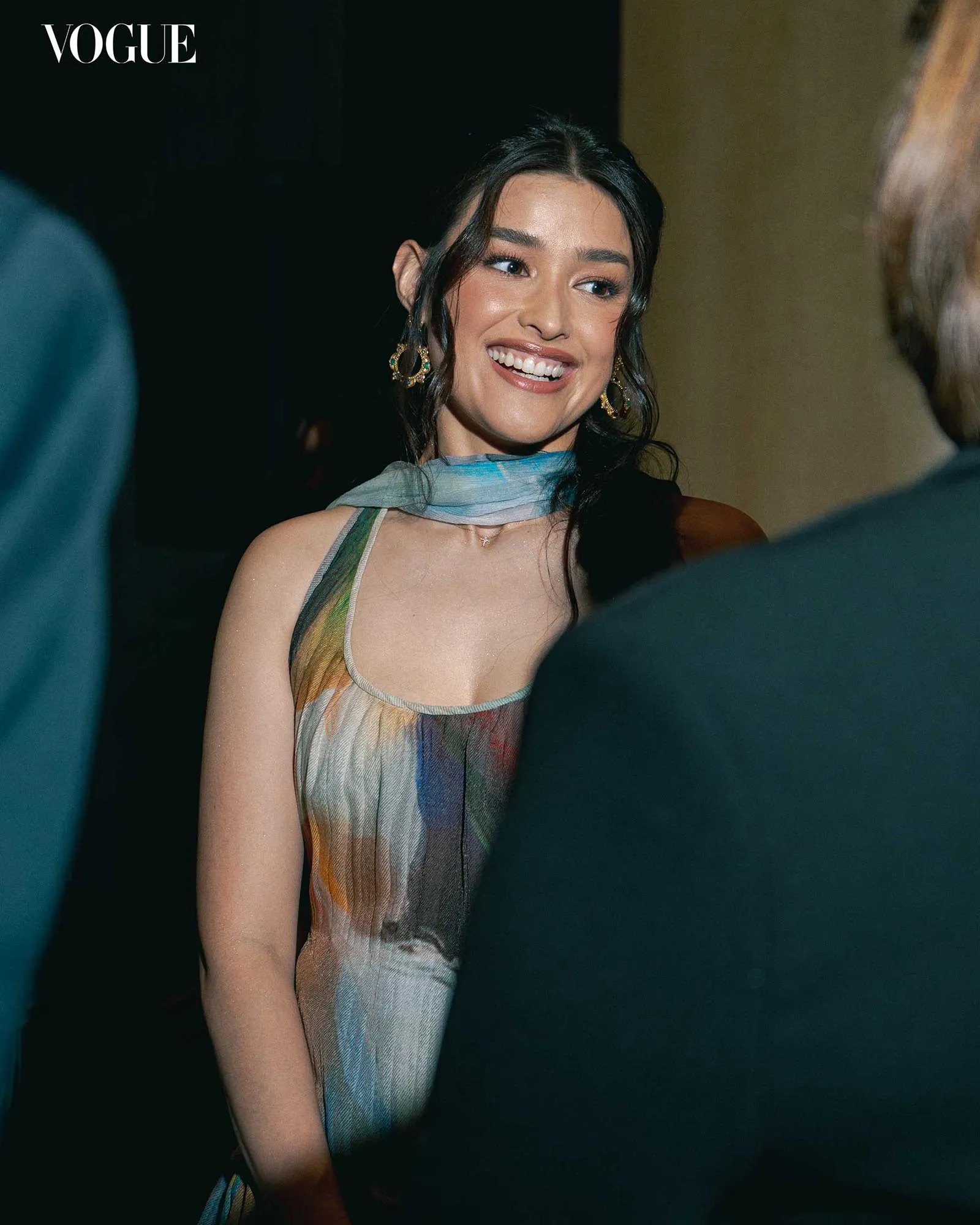 Smiling woman in a colorful striped dress with a light scarf; Vogue logo in the top-left corner.
