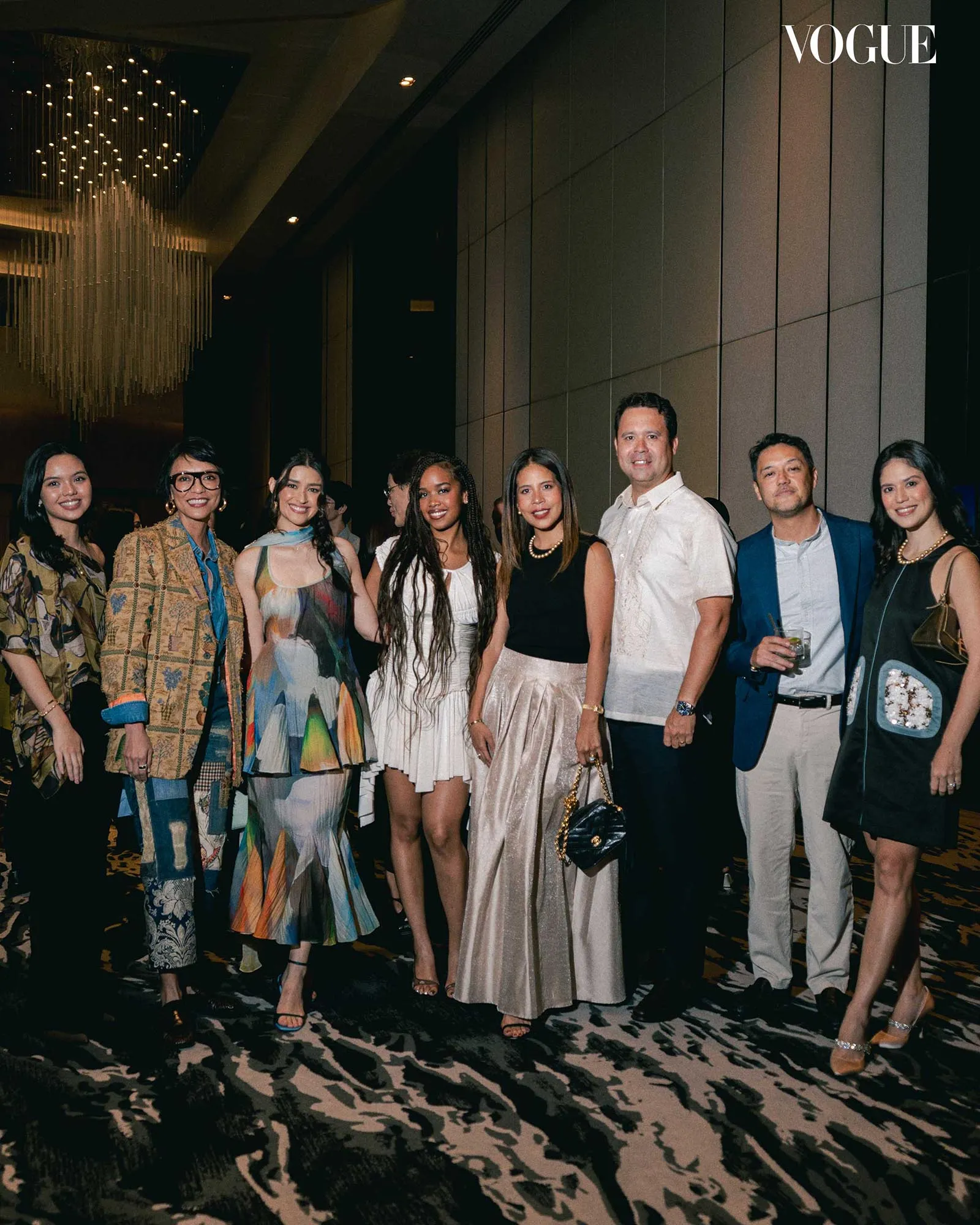 Group of fashionably dressed people posing at a Vogue event in a luxe venue, chandelier overhead.