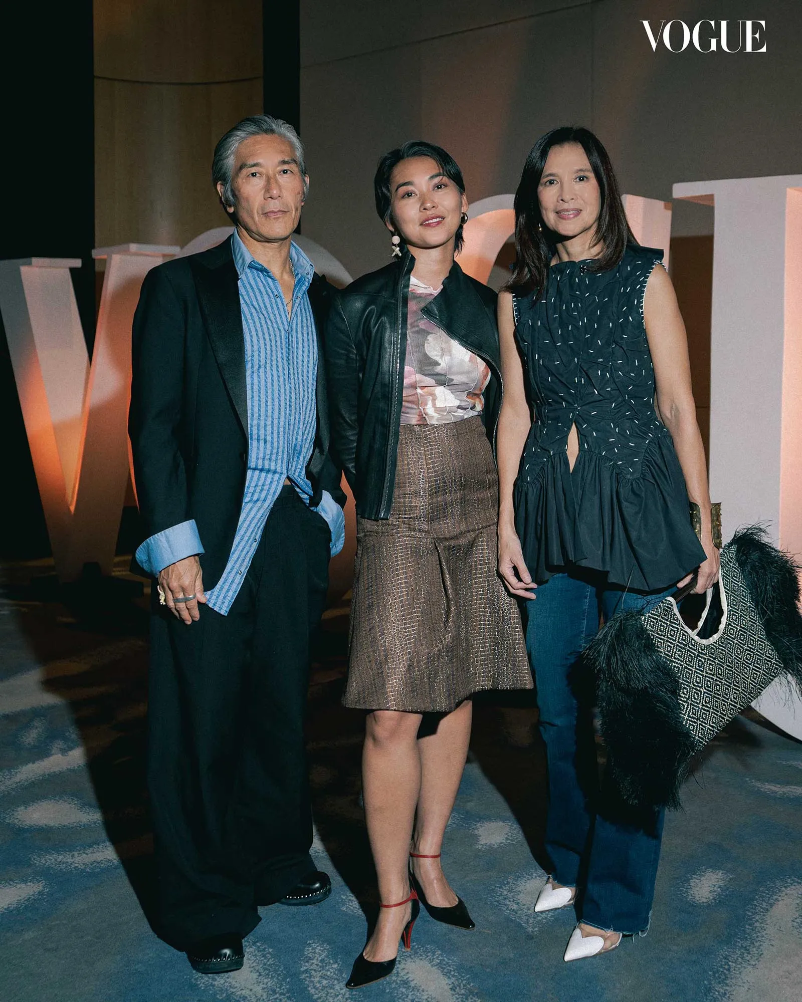 Three stylish adults posing for a Vogue event; man on the left in a blazer, woman in the center with a leather jacket and metallic skirt, woman on the right in a navy patterned dress; Vogue logo in the corner.