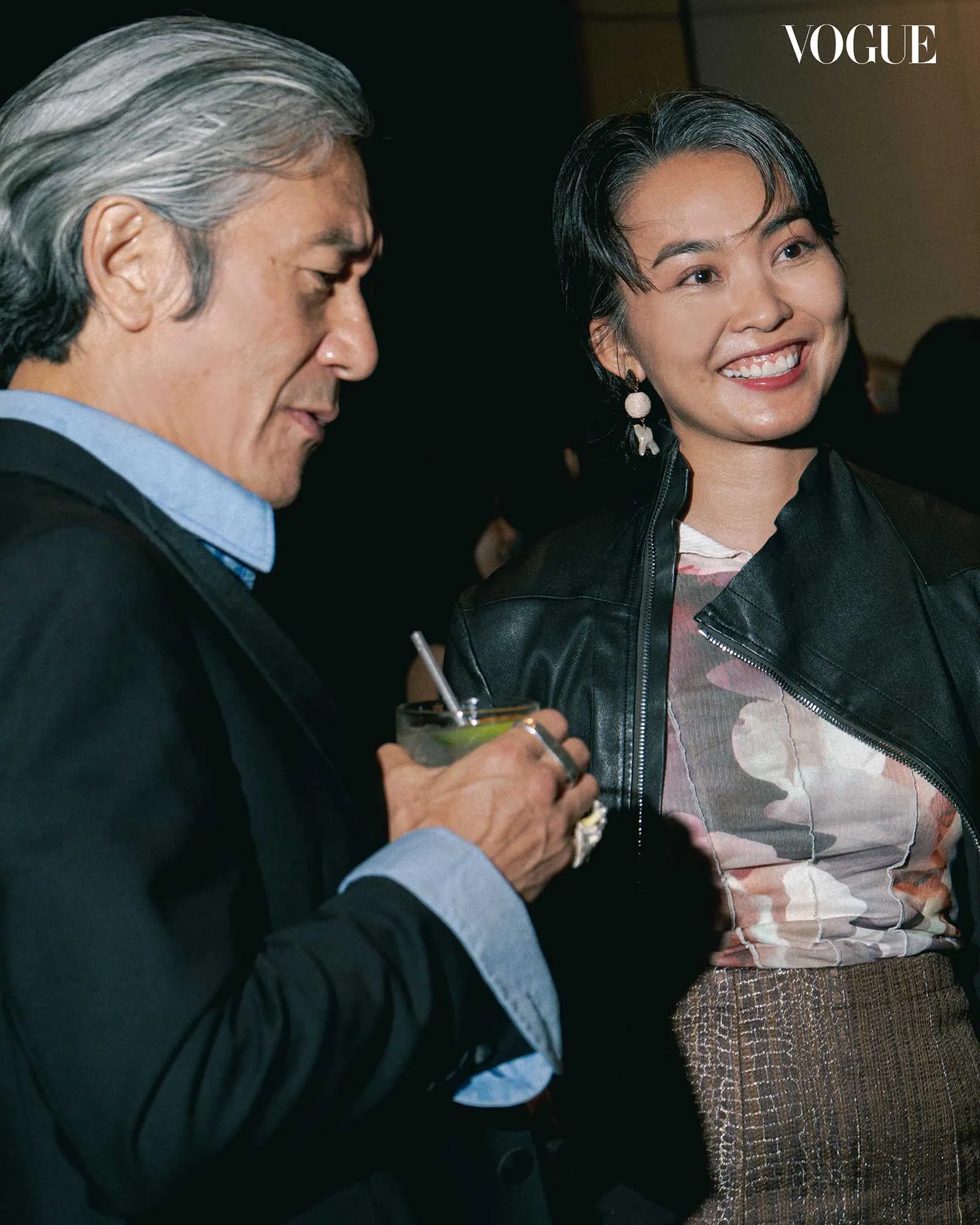Man with gray hair in a blue shirt talks to a smiling woman wearing a camouflage top and black leather jacket; he holds a drink with a straw.