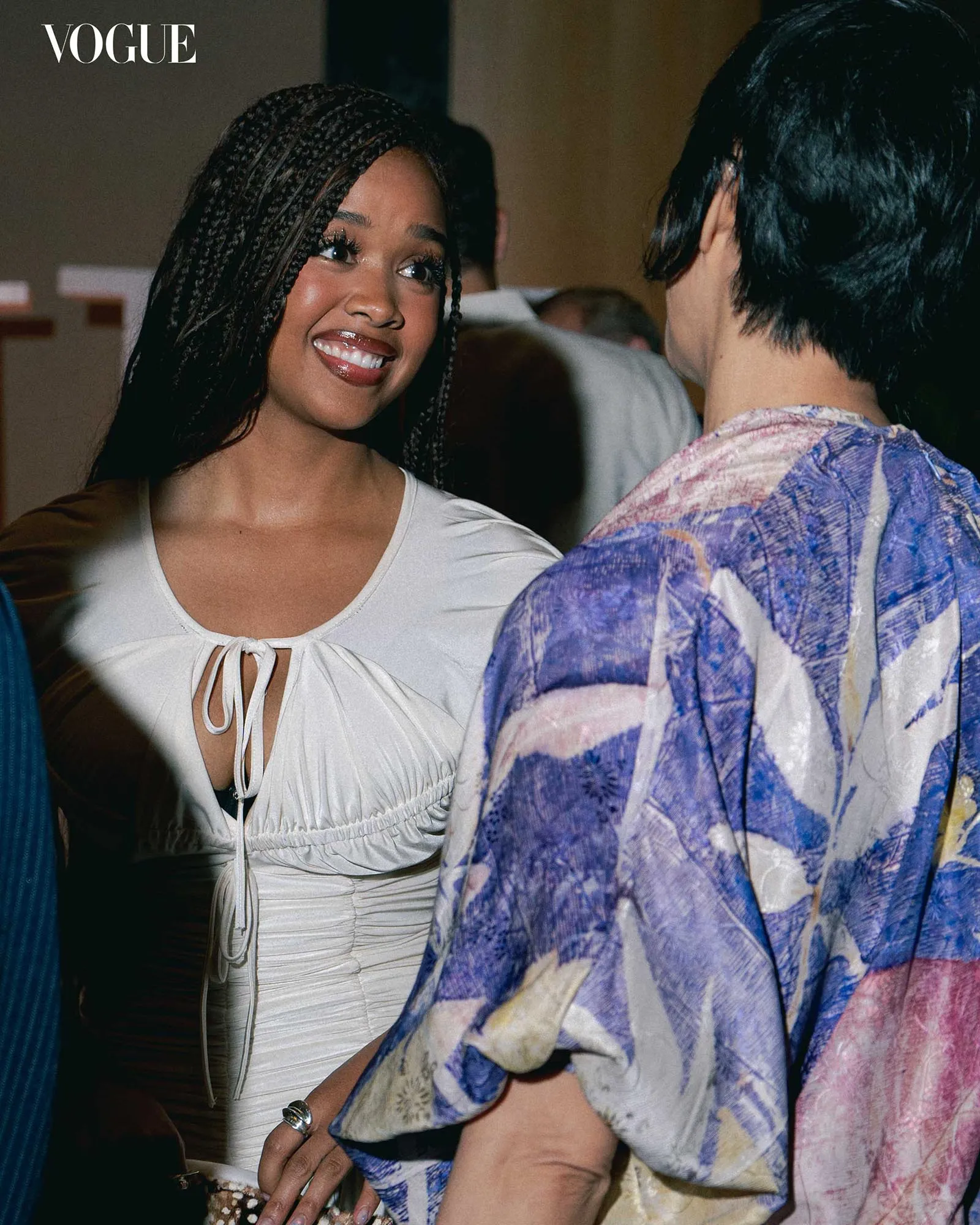 Smiling woman with braided hair in a white top talks to a person in a colorful patterned shirt at a social event; Vogue logo visible in the corner.