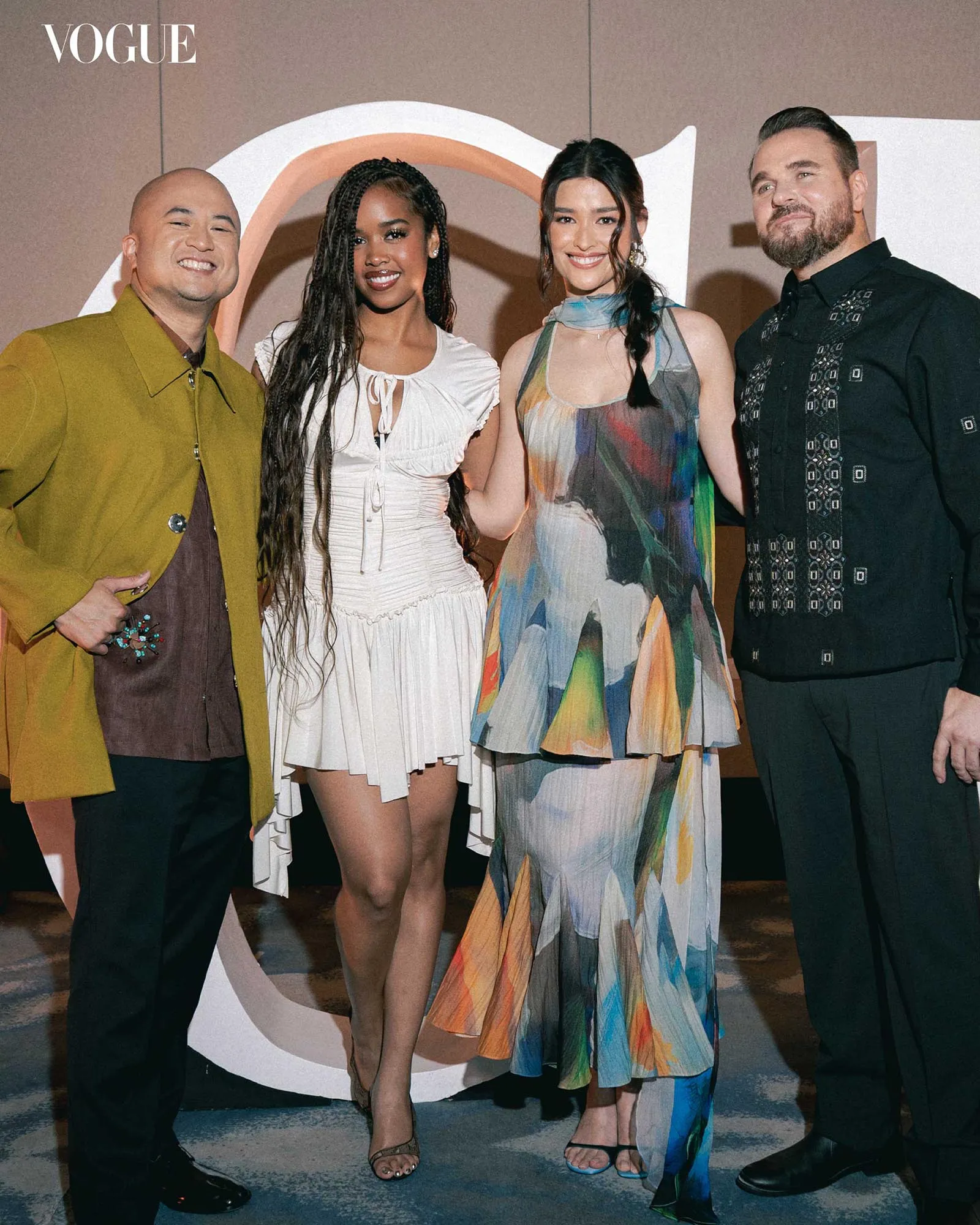 Group of four adults posing at a Vogue event, smiling for the camera in stylish outfits: mustard coat man, white dress woman, multicolor dress woman, and patterned black shirt man.