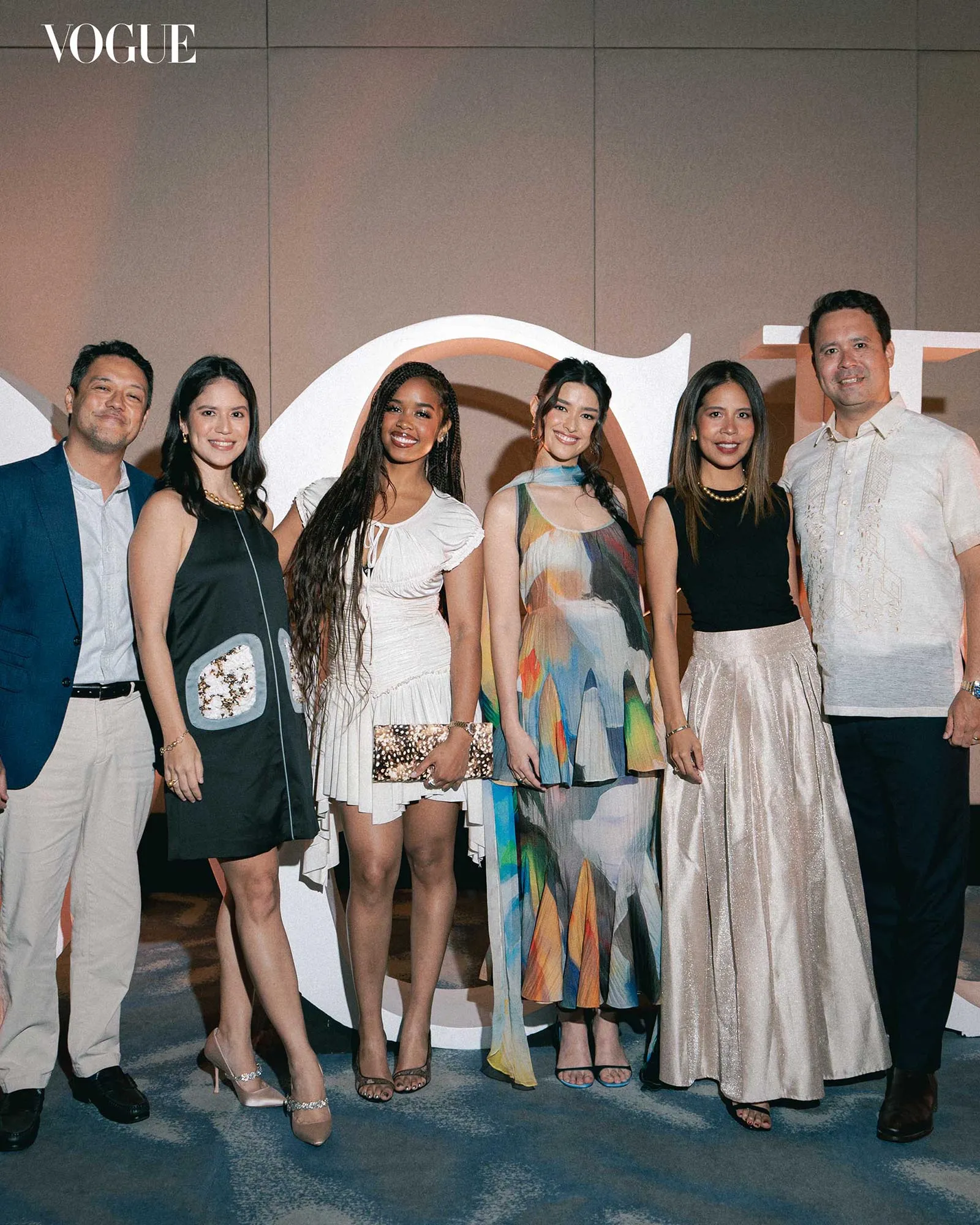 Group of six adults posing for a fashion event photo in front of a Vogue backdrop; women wear stylish dresses, men in smart outfits. Vogue logo visible in the top-left corner.