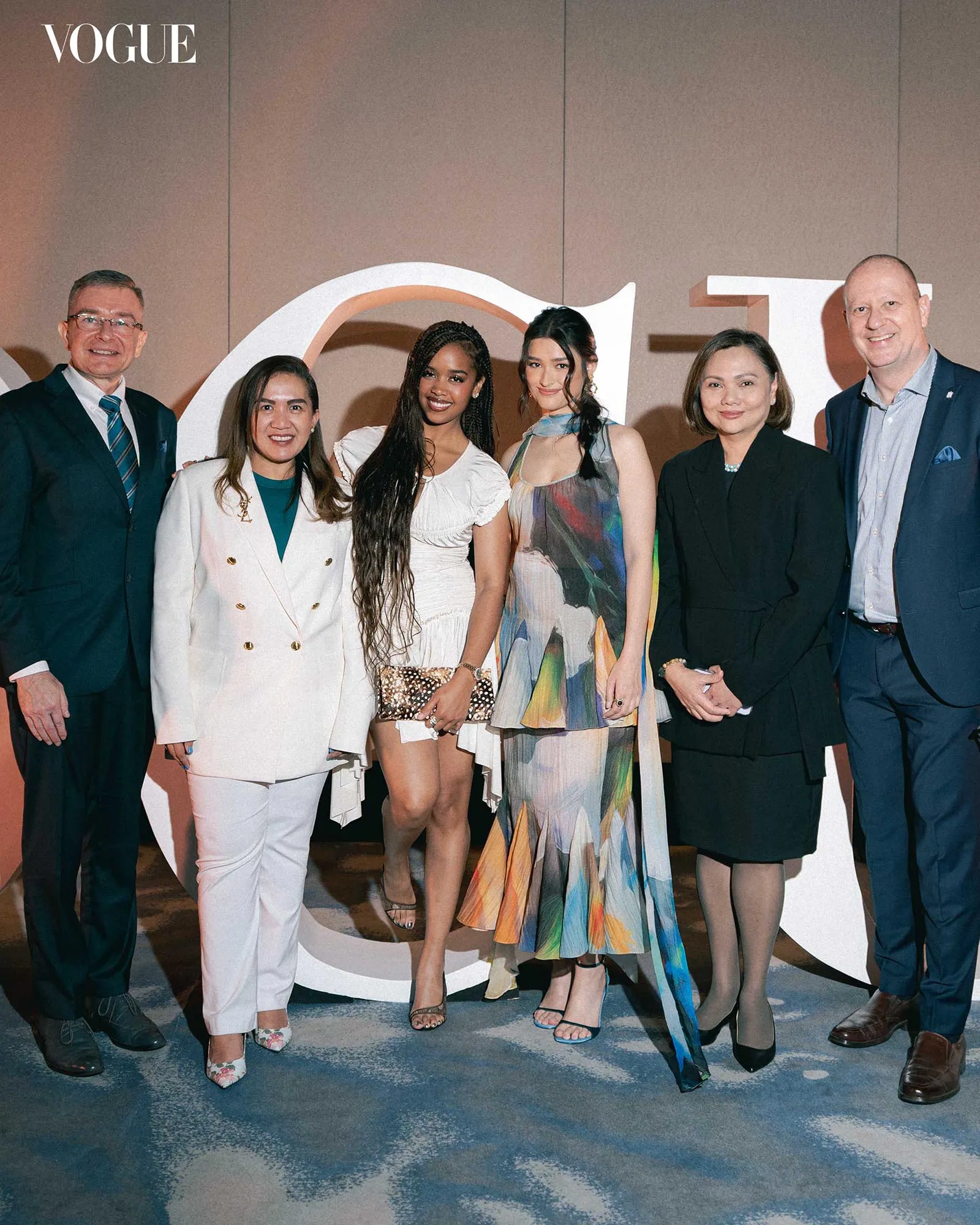 Six well-dressed individuals posing together at a Vogue event, with the Vogue logo visible in the background.