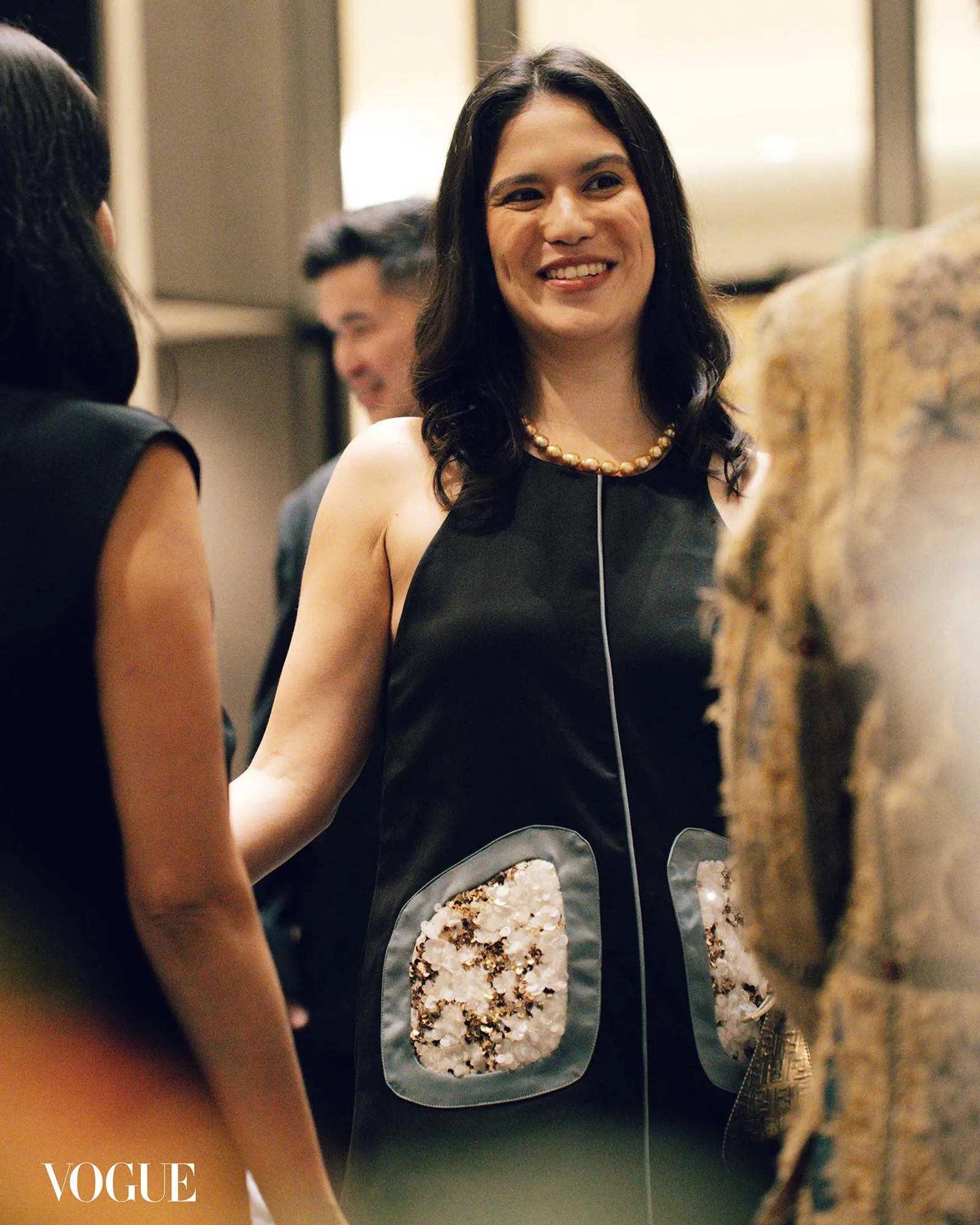 Smiling woman in a black dress with a pearl necklace at a Vogue event, blurred people in the background.
