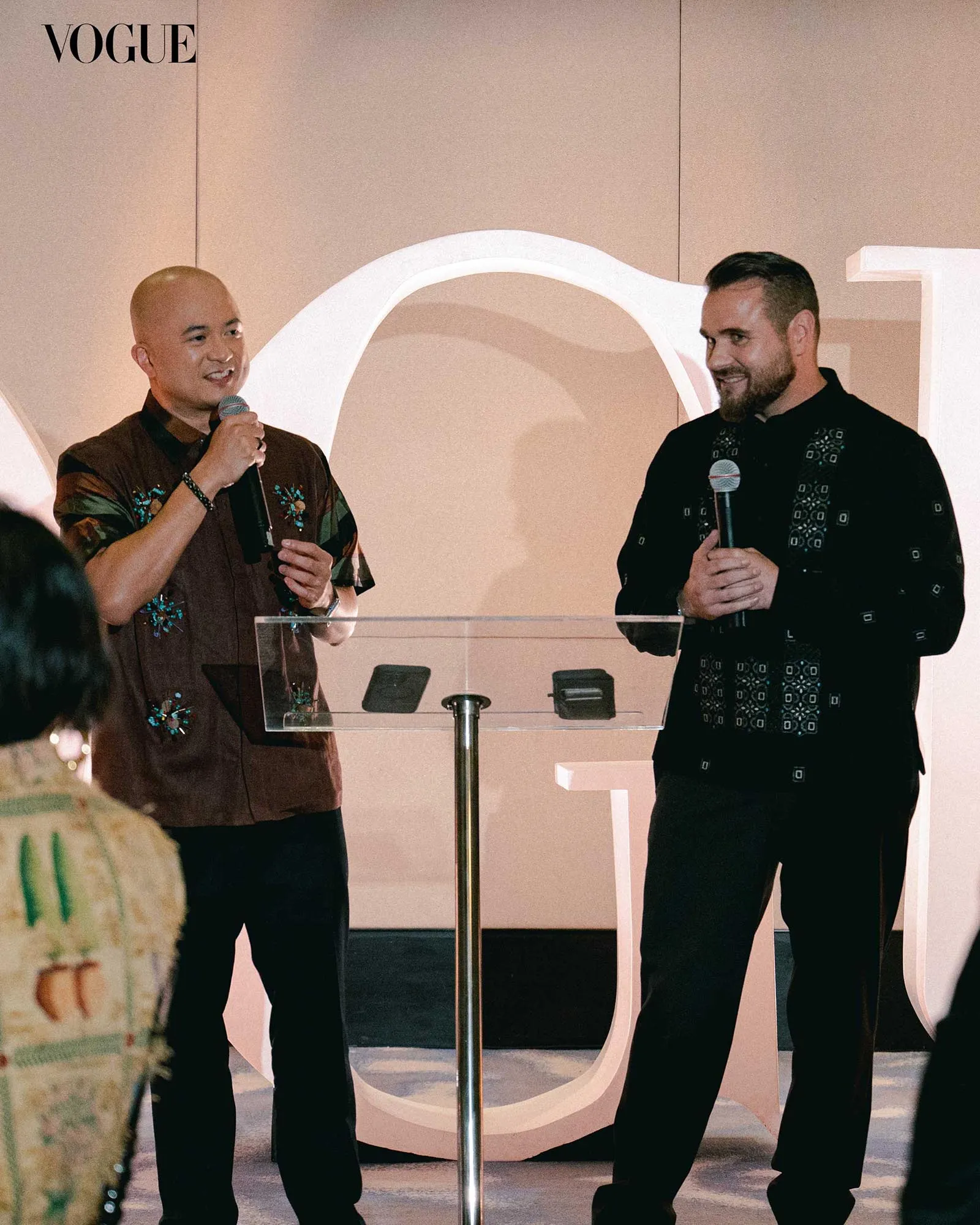 Two men speaking on a stage with microphones, standing behind a clear podium, Vogue logo in the corner, neutral backdrop.