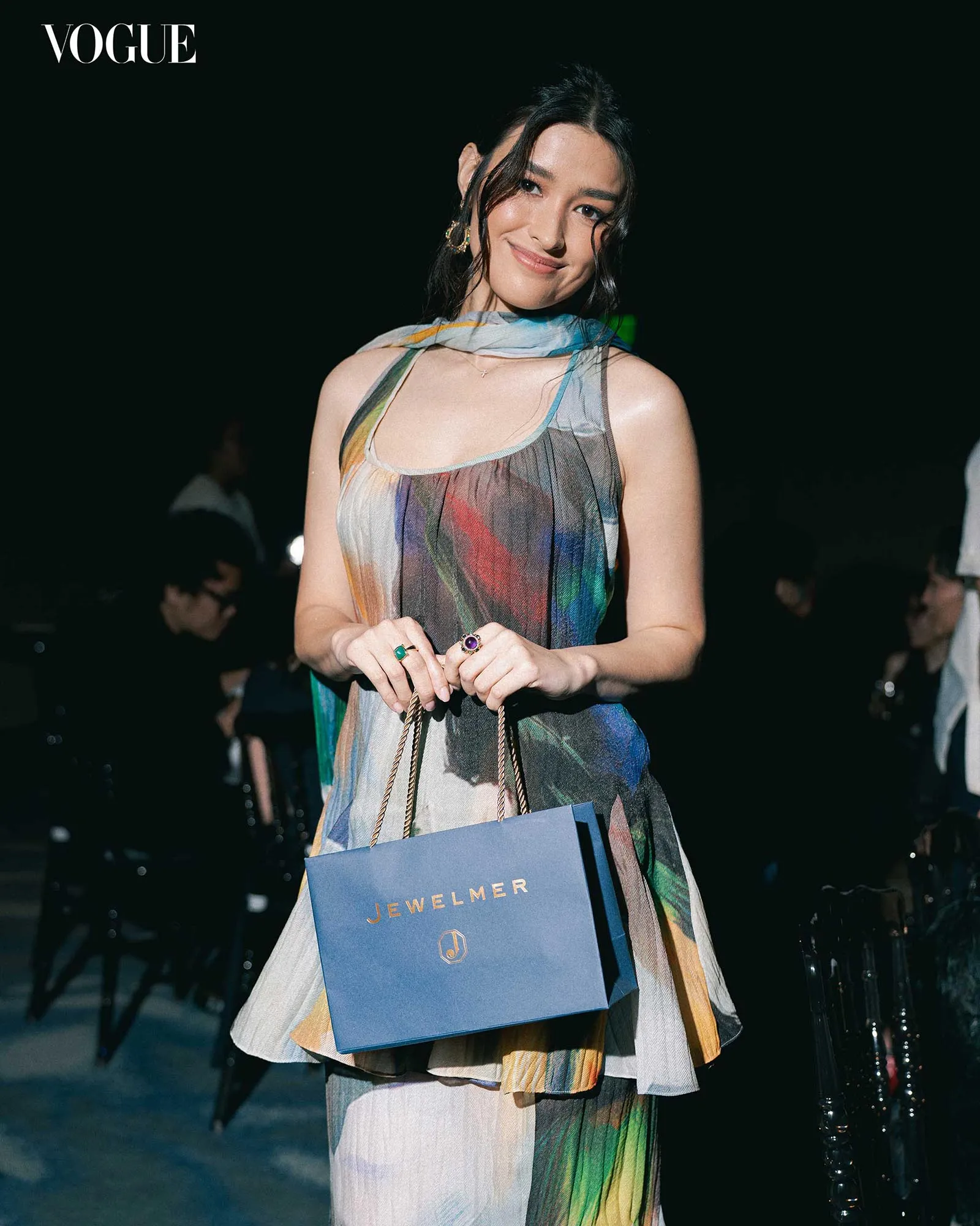 Smiling woman in a colorful sheer dress, holding a blue Jewelmer shopping bag at a Vogue event.