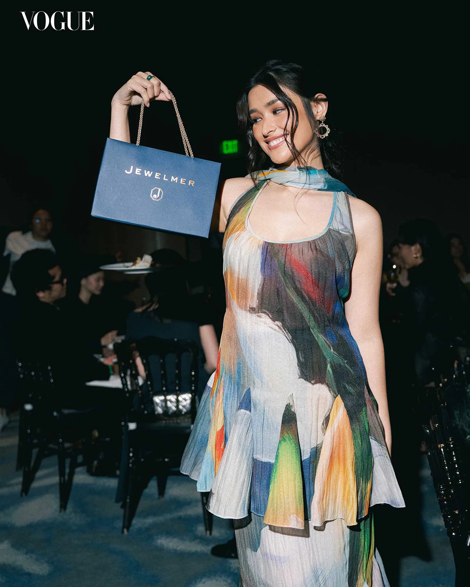 Model in a colorful dress smiles while holding a blue Jewelmer gift bag with rope handles at a Vogue event. (Fashion shoot setting)