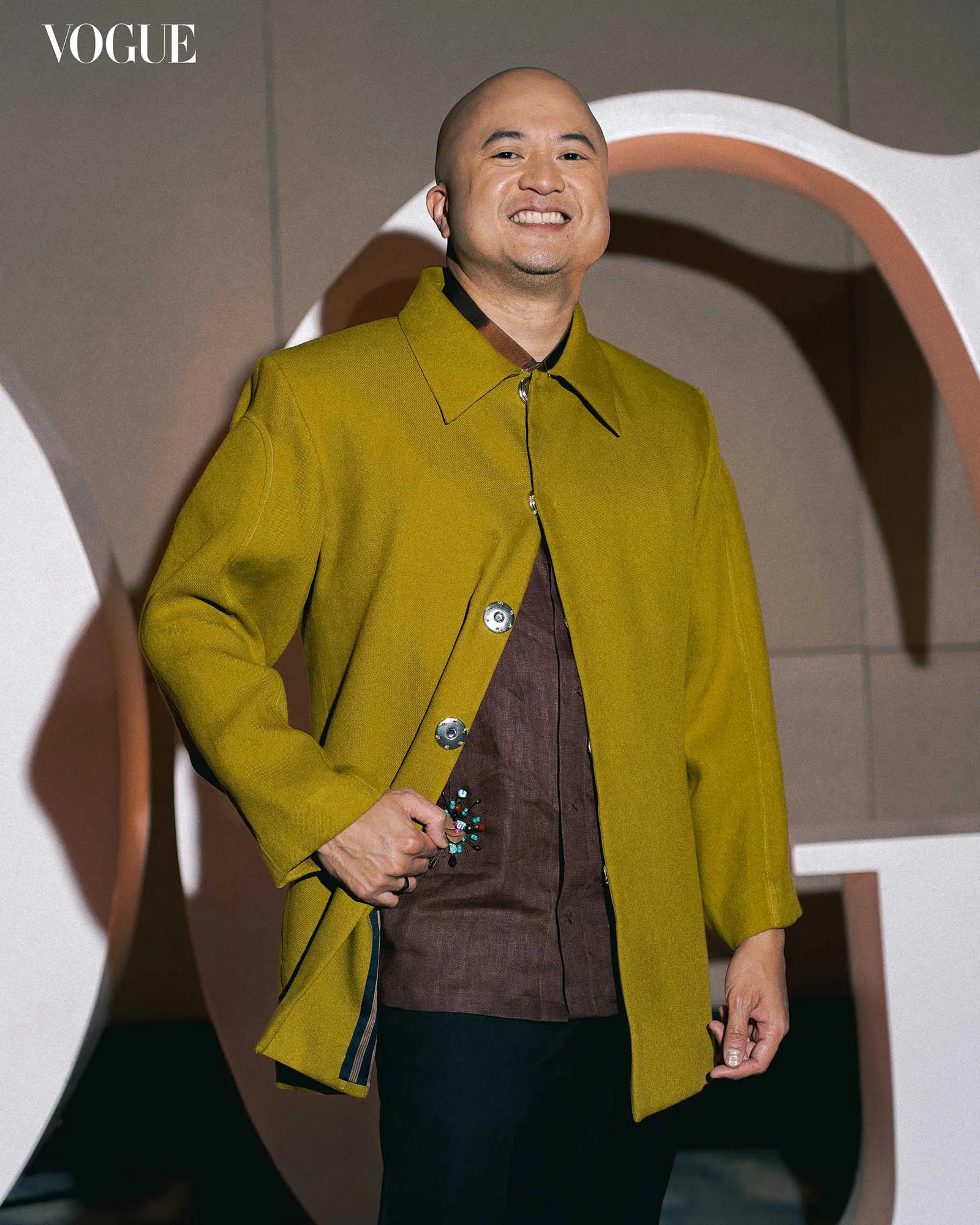 Vogue magazine lead image of a smiling person in a mustard coat, holding beaded accessory against abstract background.