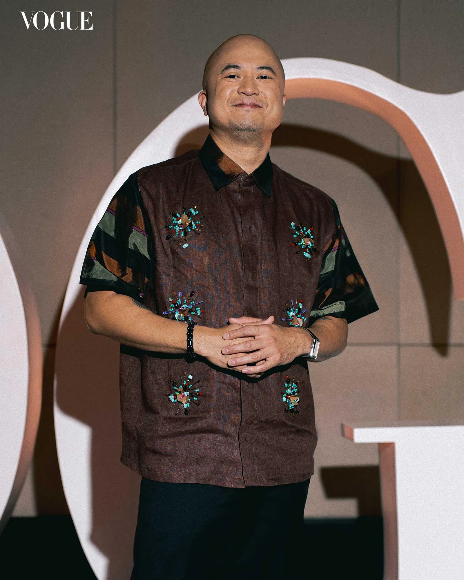 Man standing in a brown embroidered shirt with colorful beadwork, hands clasped, posing in front of a curved backdrop with Vogue logo in the corner.