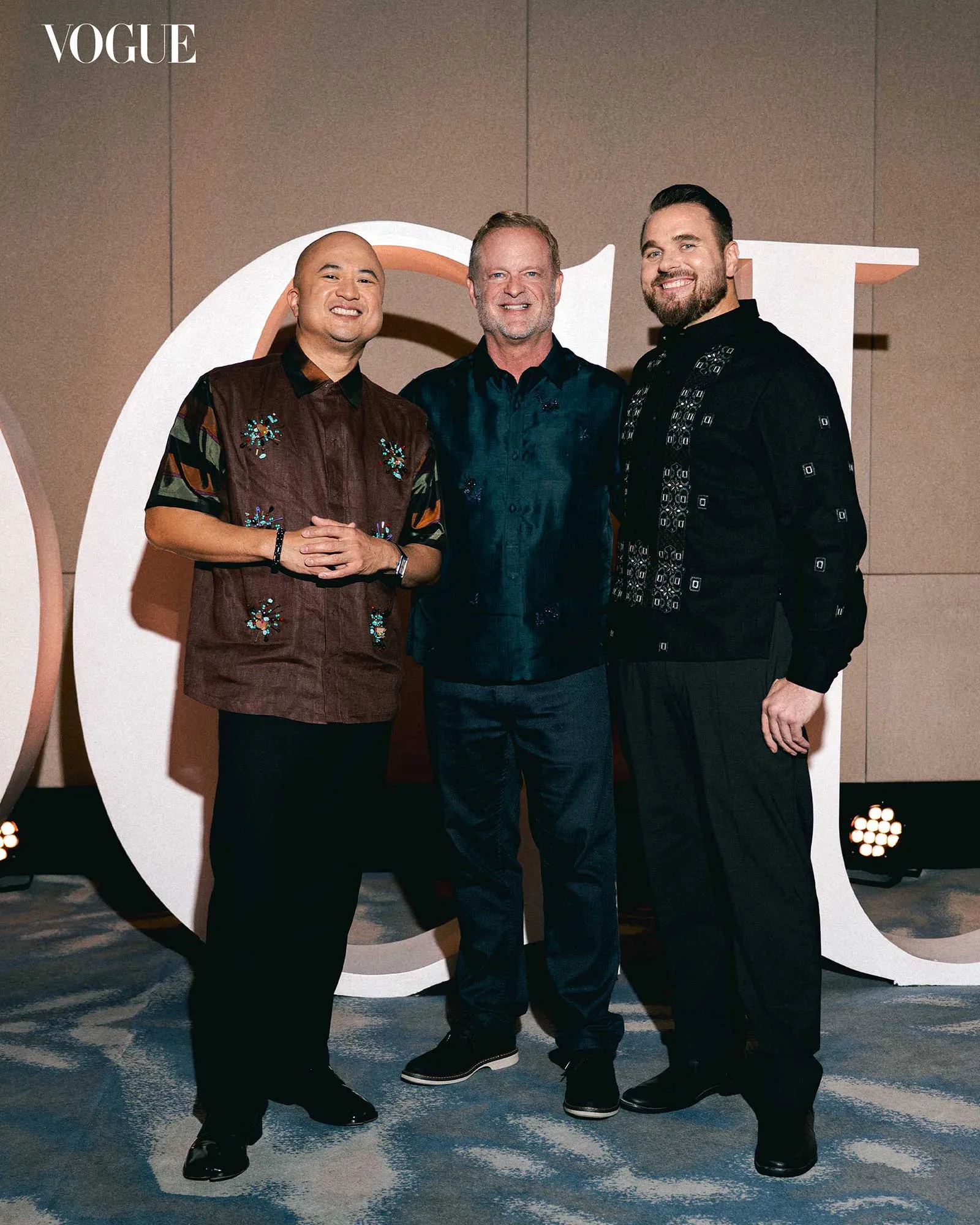 Three men in stylish outfits posing for a photo at a Vogue event with a large decorative backdrop behind them.