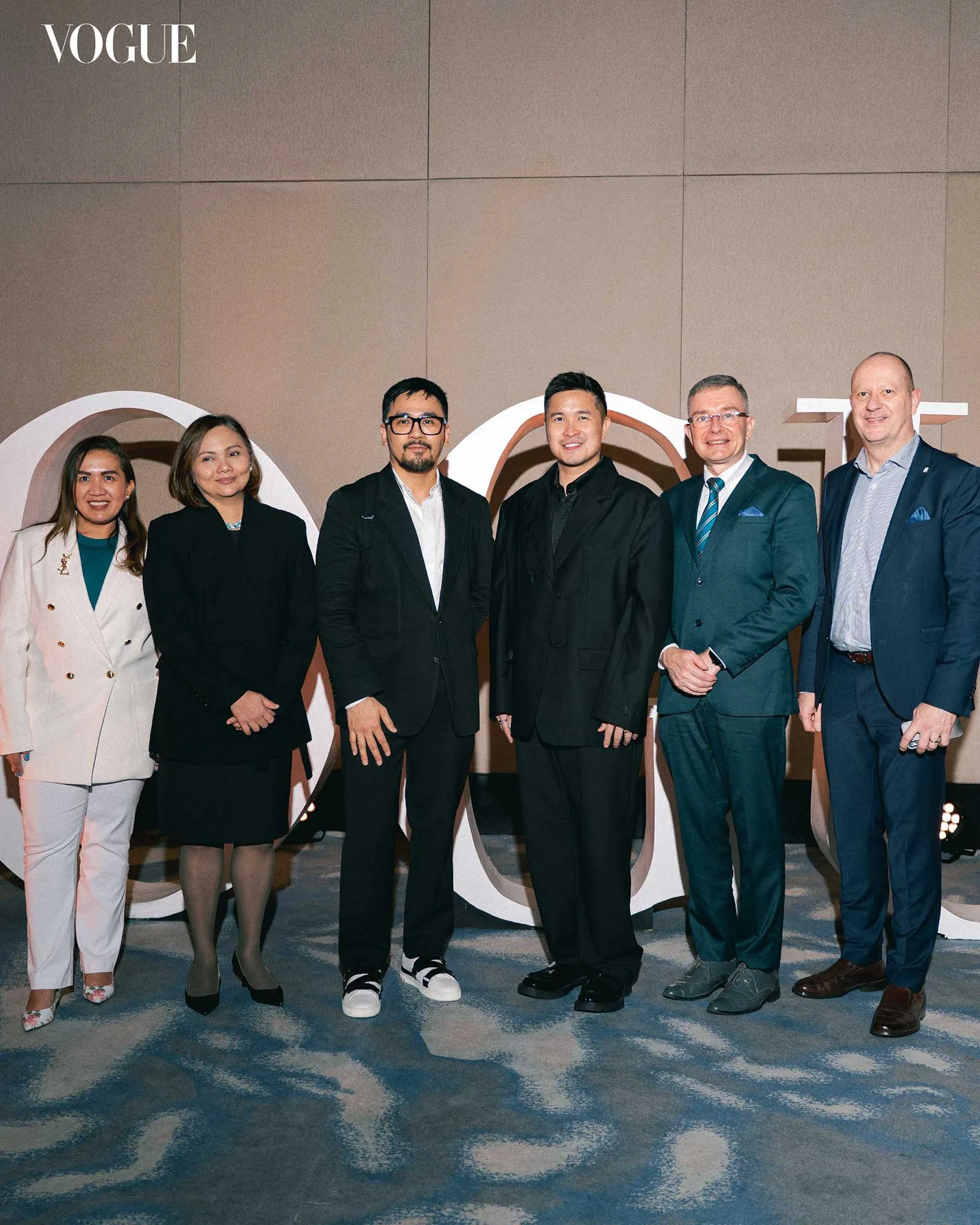Six professionals posing for a group photo at a Vogue event, in front of a backdrop with large Vogue lettering behind them.