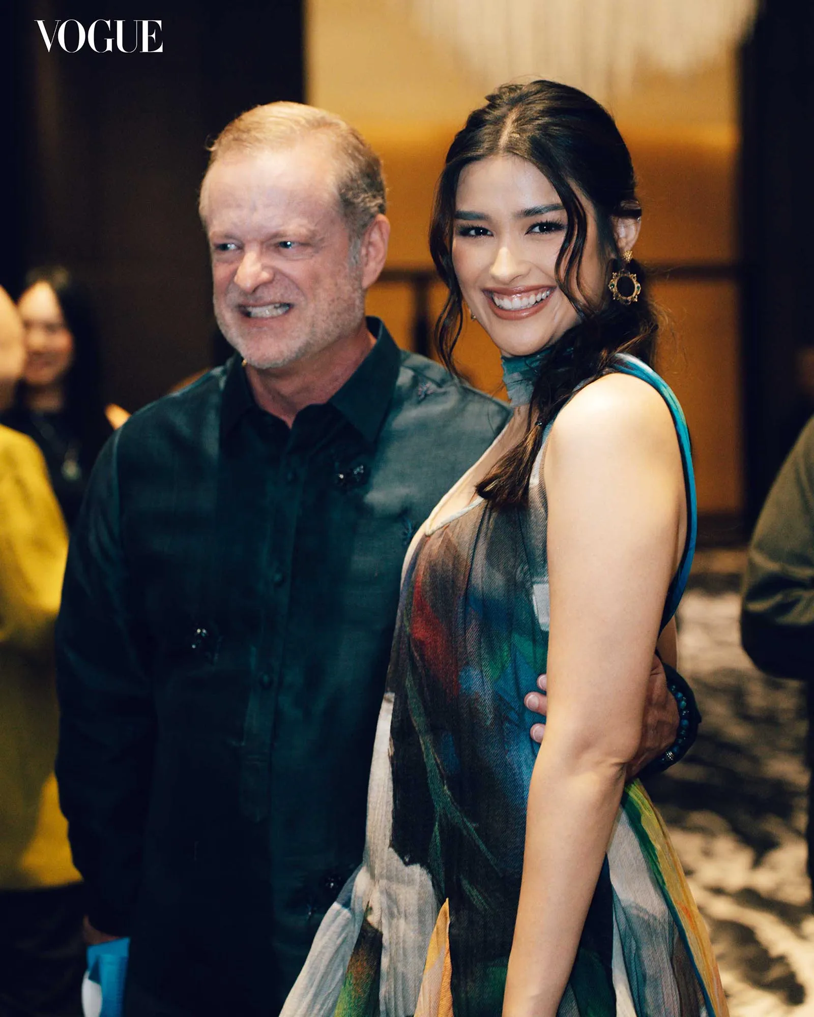 Two adults posing for a photo at a Vogue event; woman in a colorful, patterned dress with gold earrings, man in a dark shirt smile beside her.