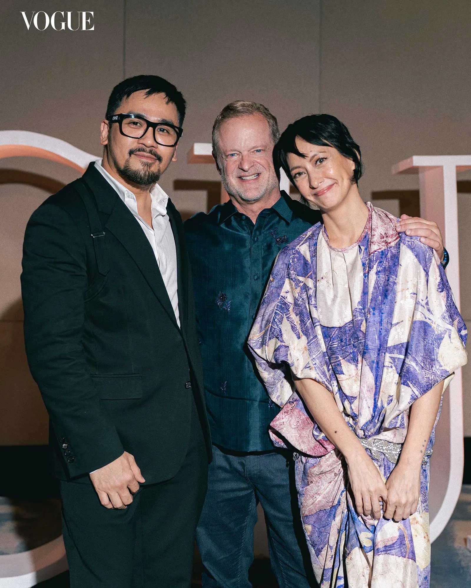 Three people pose together at a Vogue event: a man in a black suit with glasses, the smiling older man in the middle, and a woman in a purple patterned dress on the right.