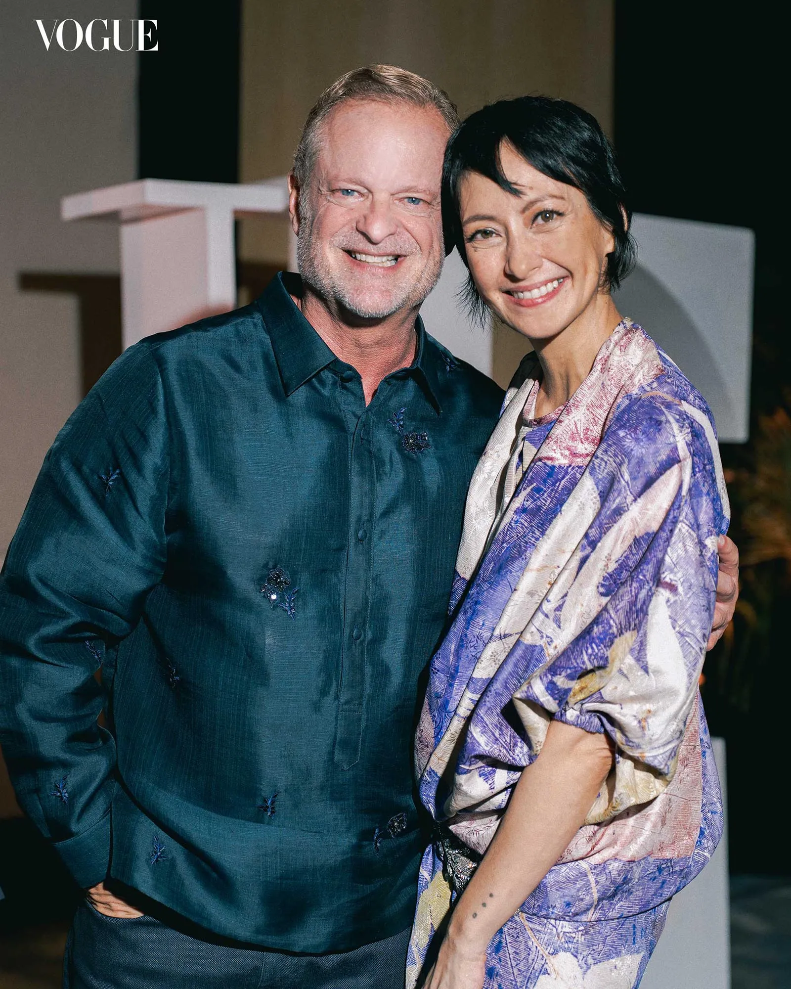 Smiling couple posing for a Vogue photo; man in teal shirt and woman in a purple scarf.