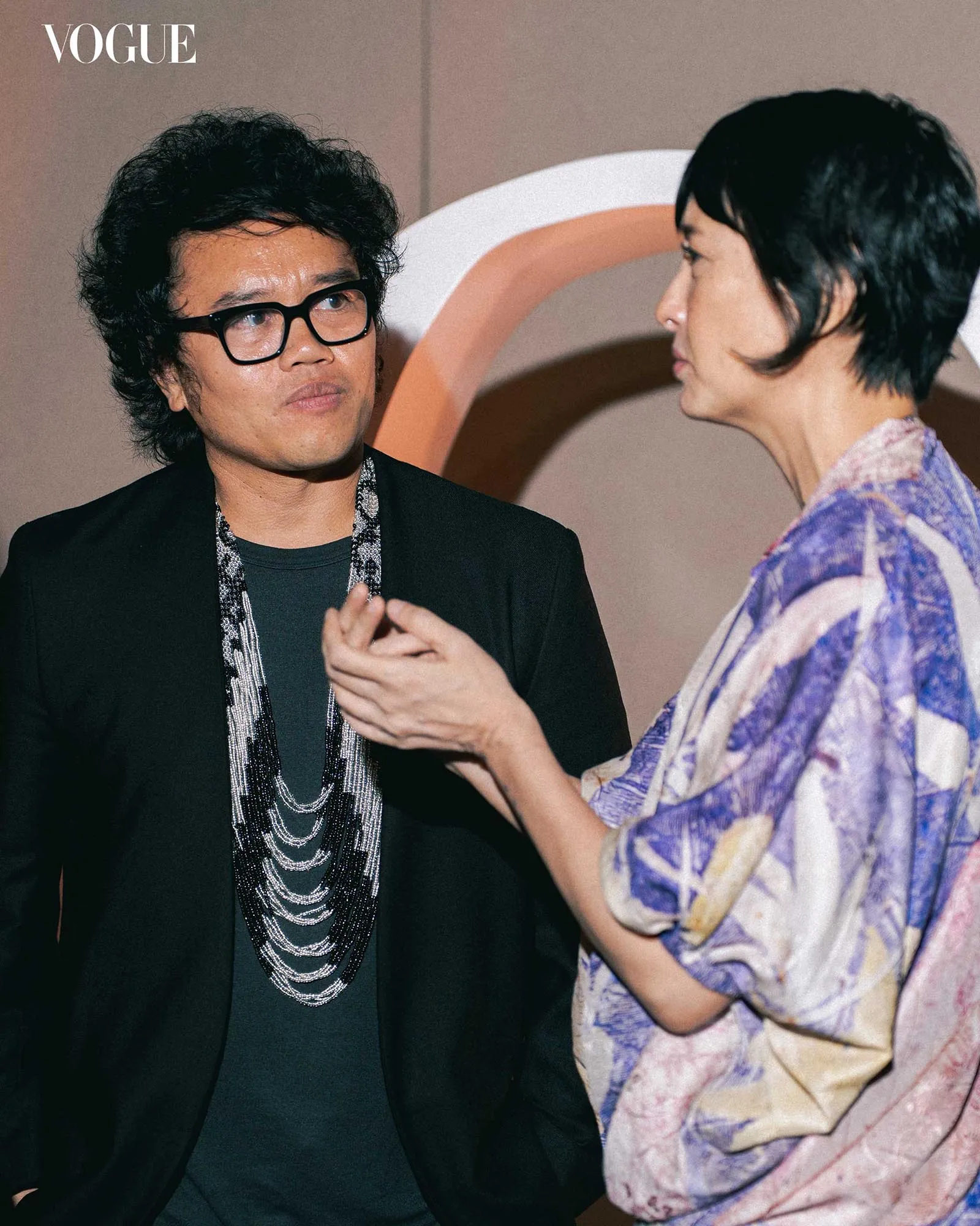 Two people in conversation at a Vogue event: a man with black-framed glasses and layered silver necklaces wearing a black blazer, facing a woman in a purple patterned outfit.
