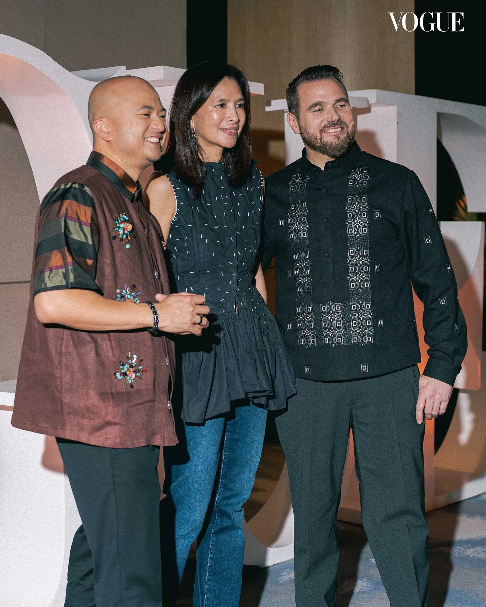 Three adults posing and smiling at a Vogue event: left man in embroidered brown vest, center woman in a dark dress, right man in a black embroidered shirt.