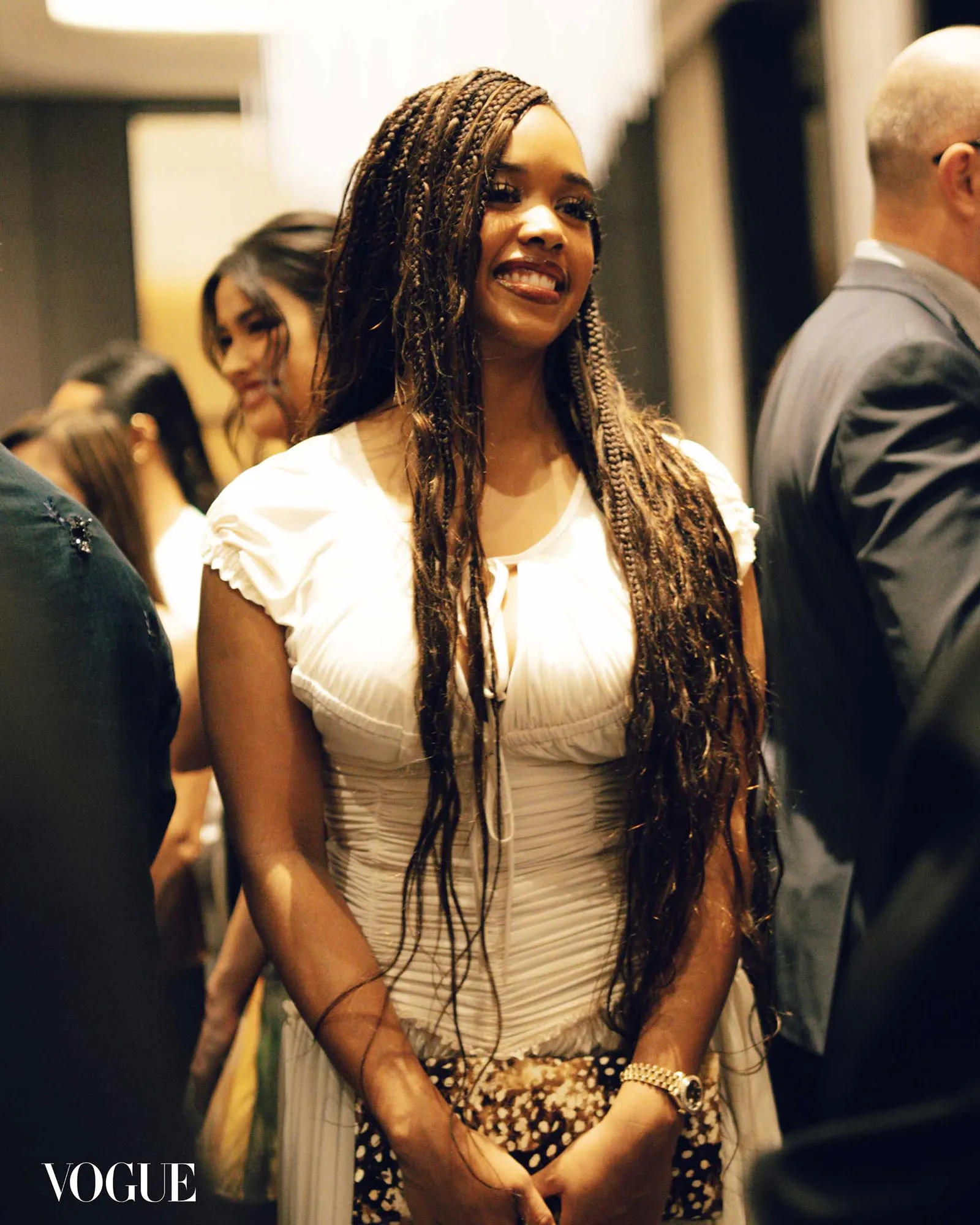 Smiling woman with long box braids in a white dress at a fashion event; Vogue logo in corner.