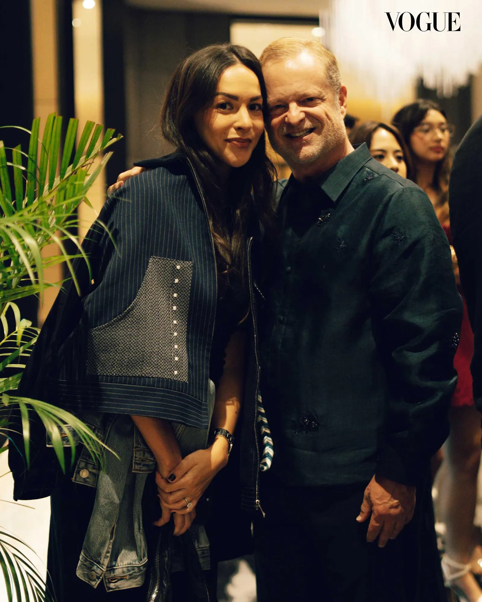 Two people stand close together, smiling for a photo at a Vogue event, with a plant in the foreground and the Vogue logo in the corner.