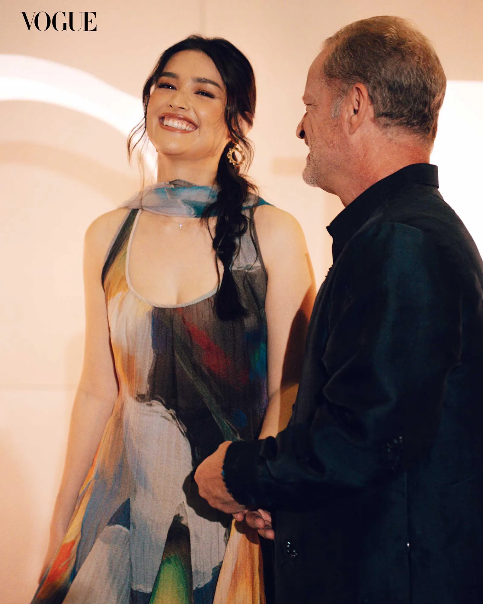 Smiling woman in a colorful dress stands beside a man in a dark shirt; Vogue logo visible in the upper left corner.