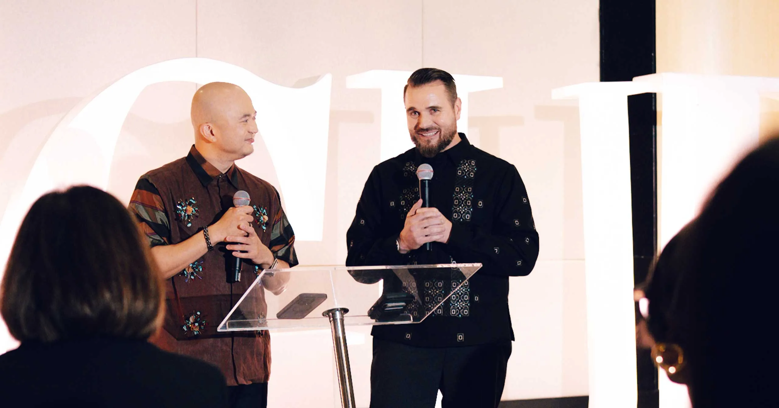 Two men on a stage speak into handheld microphones at a clear podium, engaging an audience.