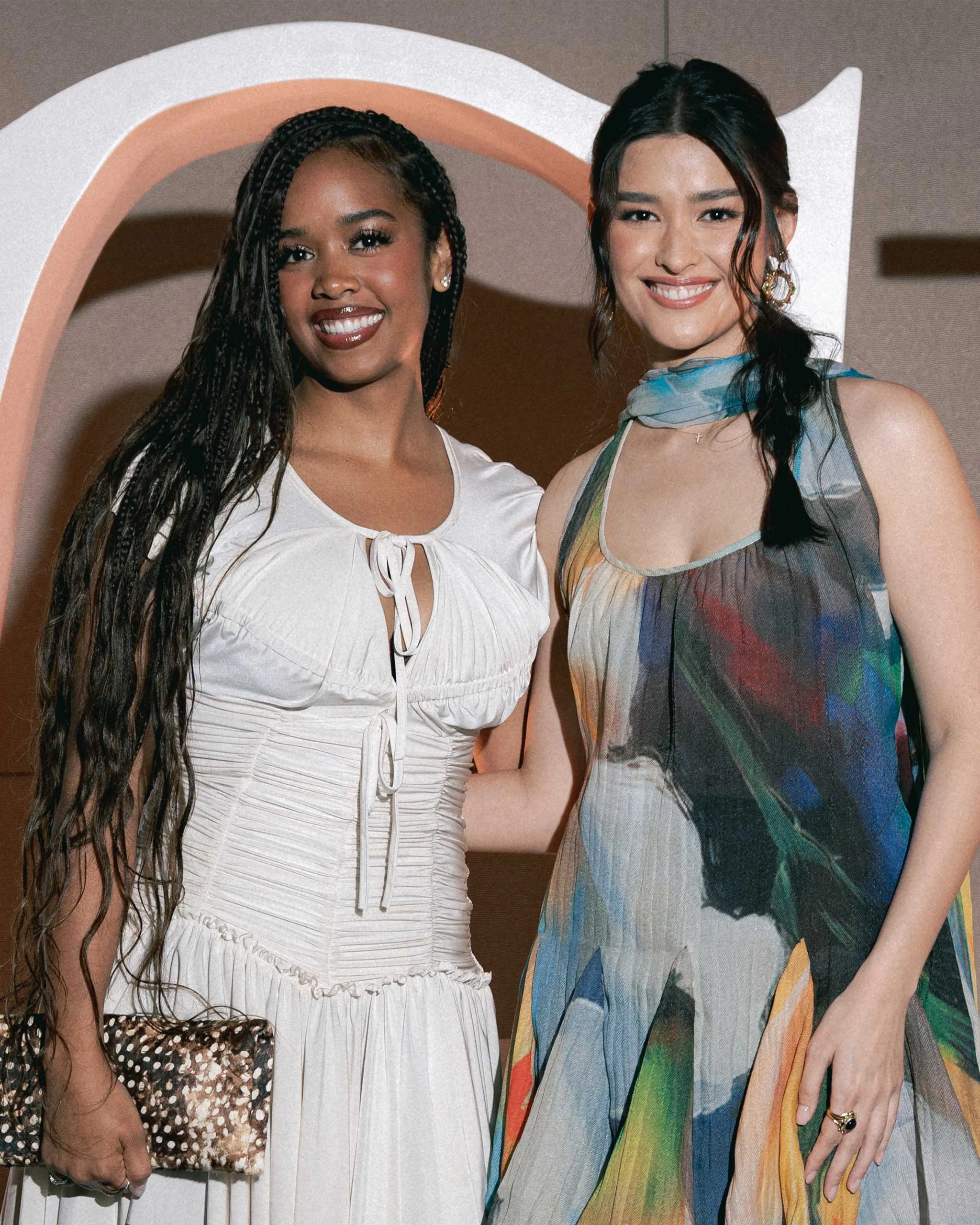 Two women pose together and smile at a formal event; one wears a white, pleated dress with long braids, the other wears a multicolored sheer dress and scarf.