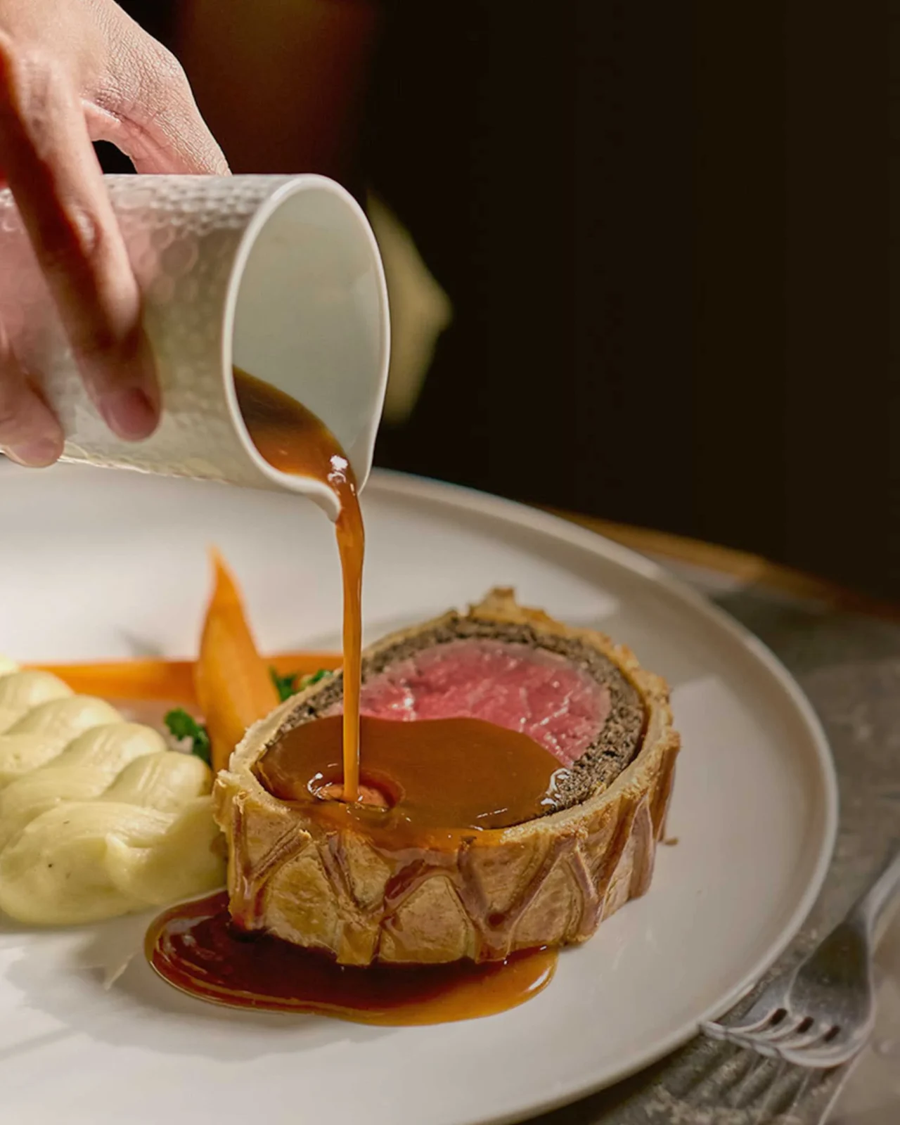 Beef Wellington on a plate with carrot puree and piped mashed potatoes, as brown sauce is being poured over the steak slices.