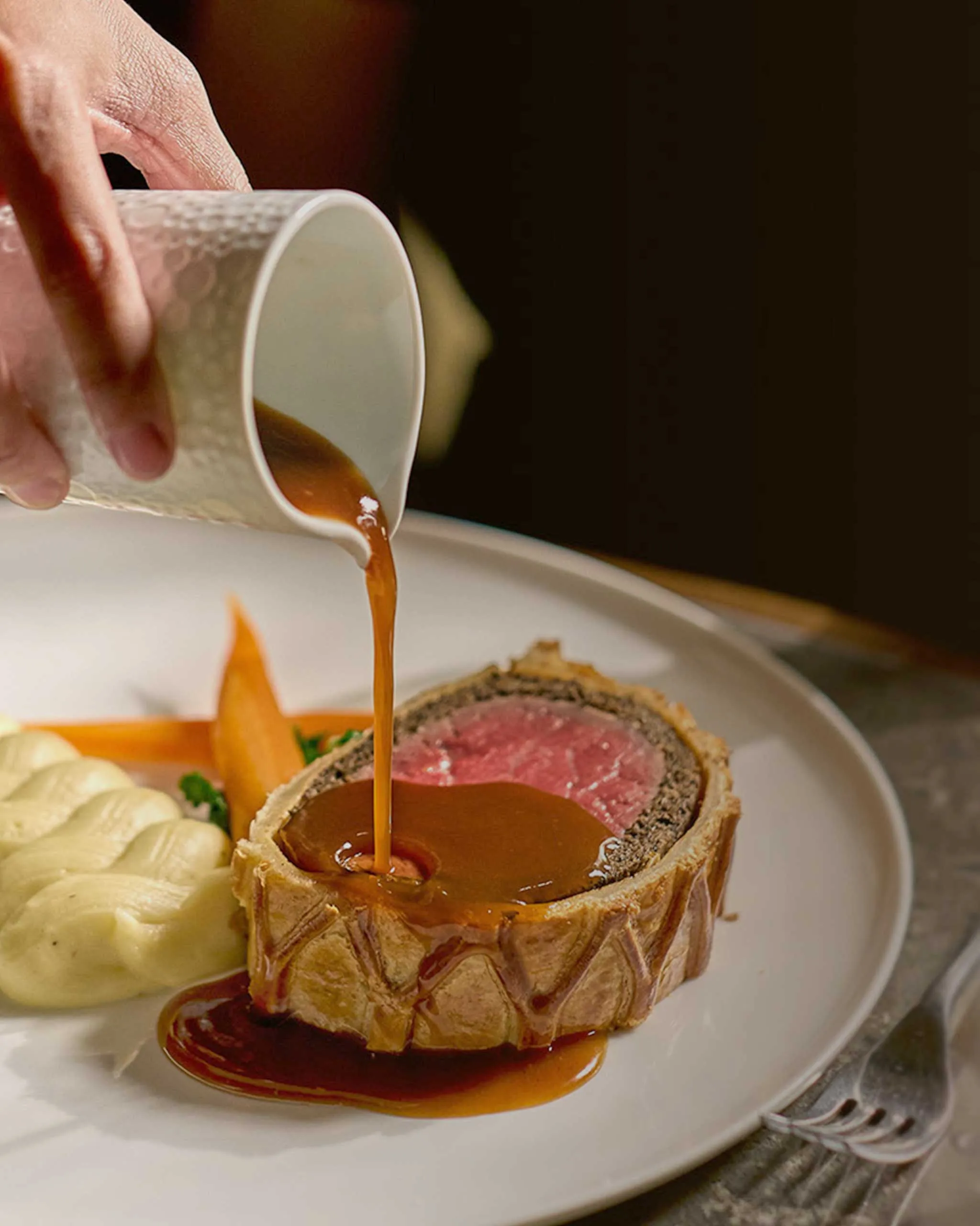Pouring brown gravy over a beef wellington half on a plate with mashed potatoes nearby and an orange carrot garnish in the background.