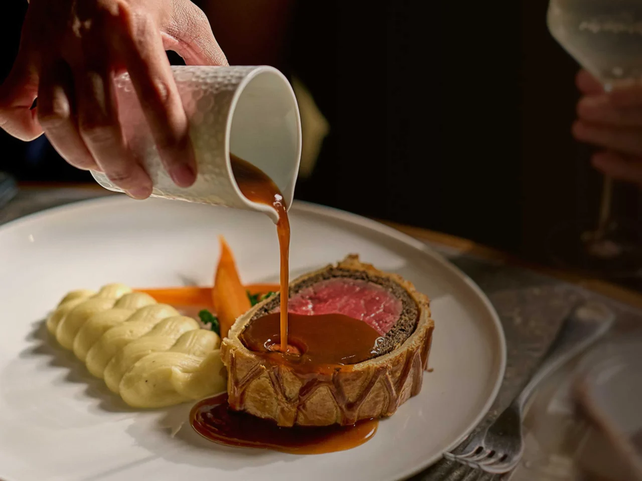 Beef Wellington on a plate with carrot puree and piped mashed potatoes, as brown sauce is being poured over the steak slices.
