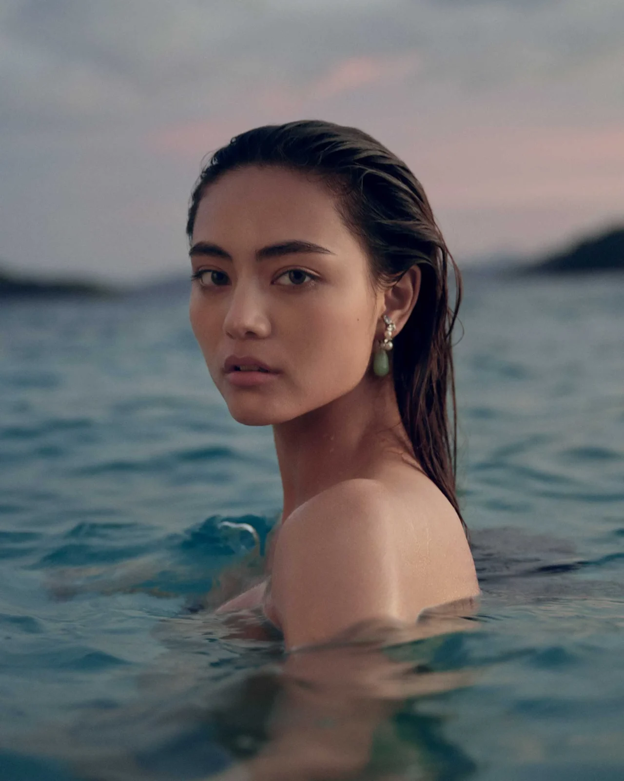 Close-up of a young woman with wet hair, wearing pearl earrings, by the ocean and looking toward the camera.