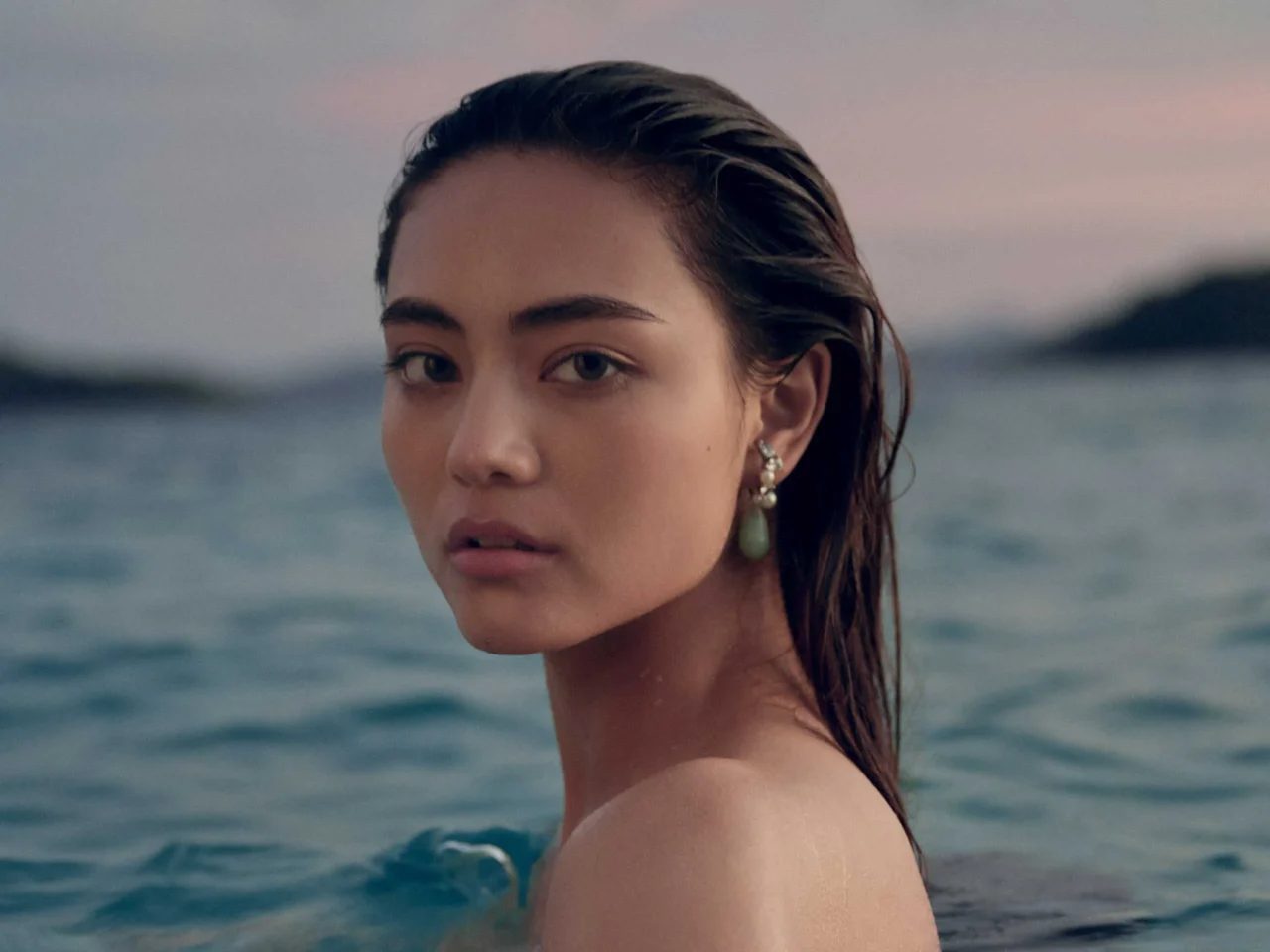 Close-up of a young woman with wet hair, wearing pearl earrings, by the ocean and looking toward the camera.