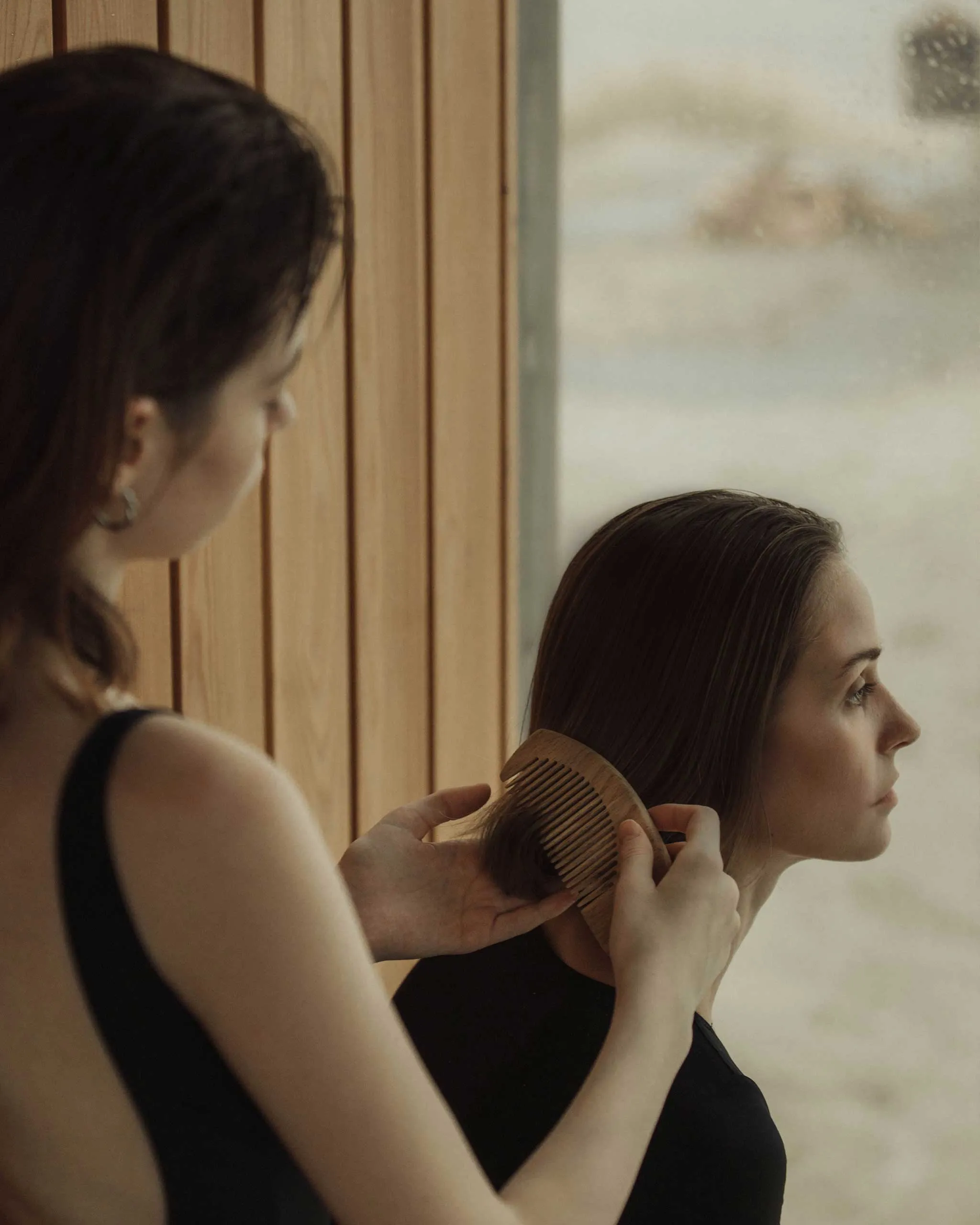 Two women in a hair salon; a stylist combs the client's hair with a wide-tooth comb near a wooden wall.