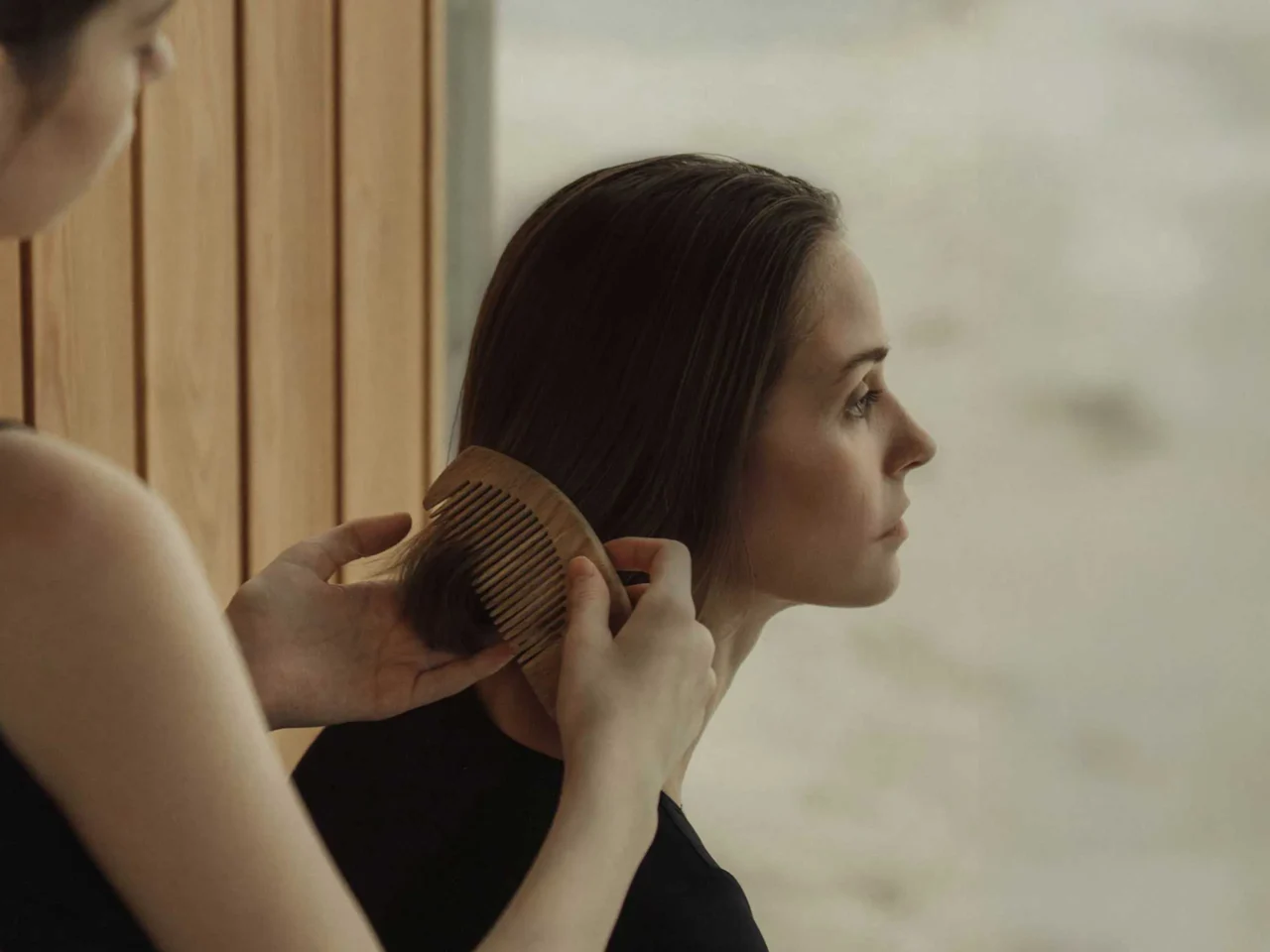 Two people brushing hair: a woman with a wooden comb trims the other’s long brown hair in a warm room with wooden panels behind them.